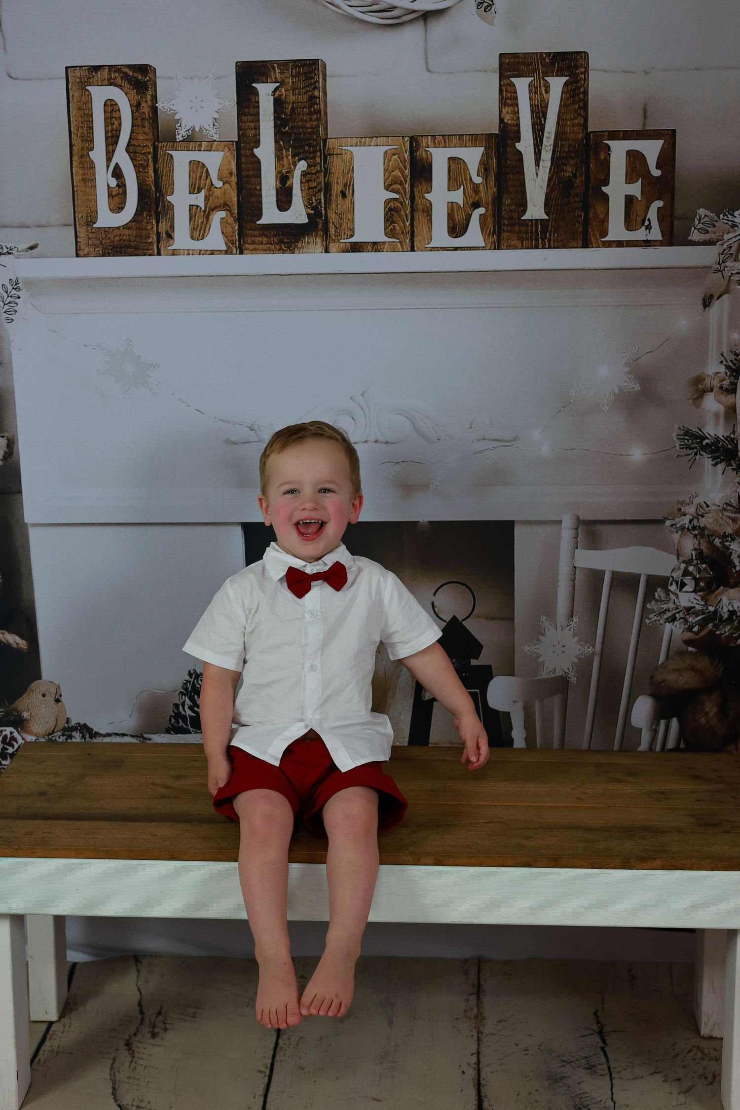 A white and wooden holiday setup features rustic 'BELIEVE' letters above a fireplace mantel and a wooden bench.