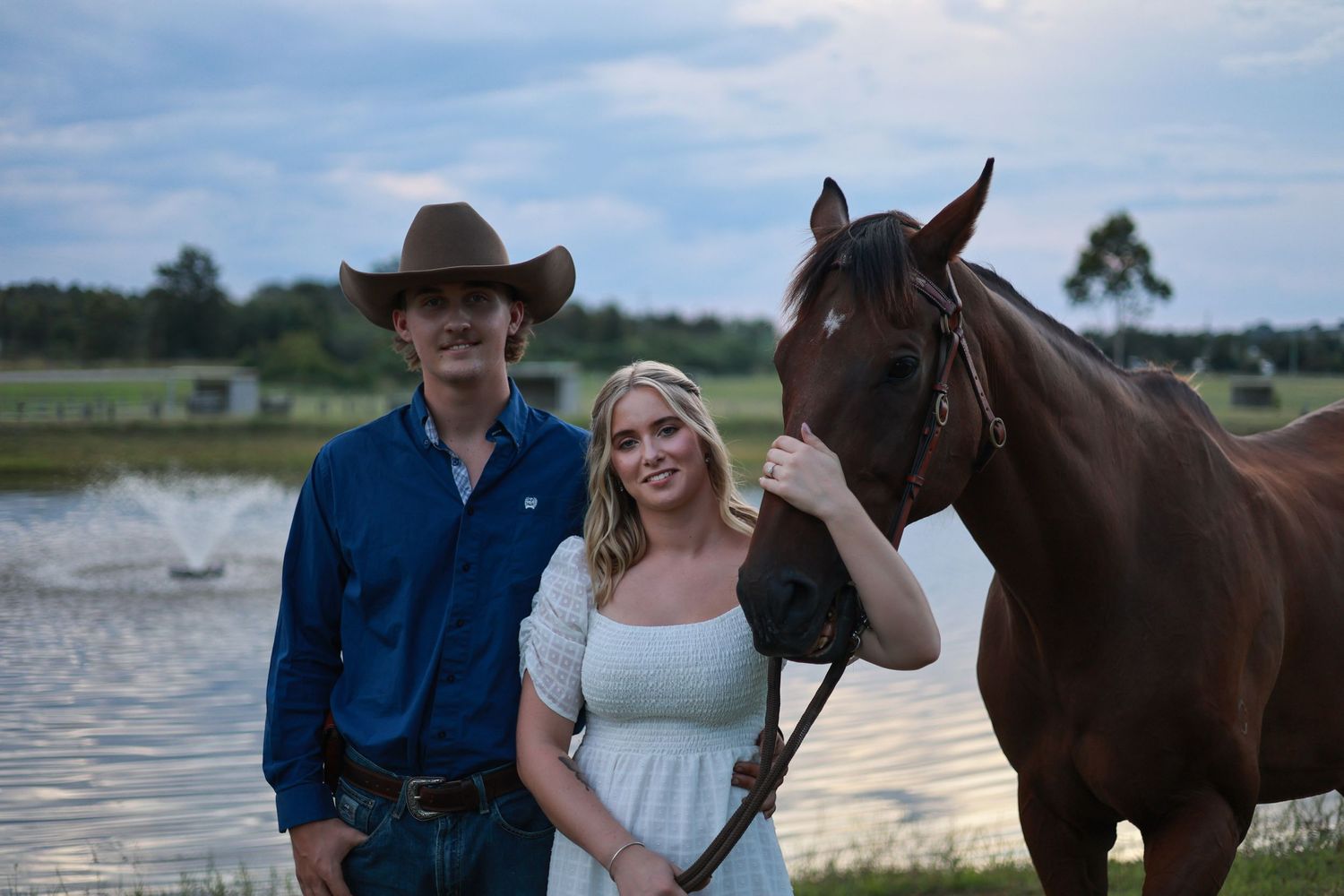 Two people stand alongside a dark brown horse near a lake at dusk.