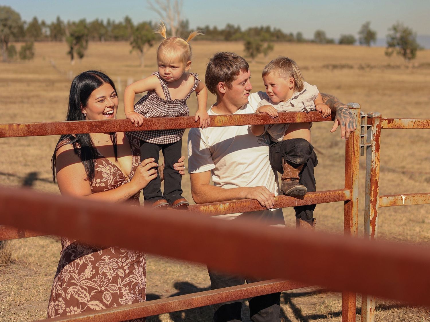 A happy family with two toddlers poses by a rustic fence in a dry rural field on a sunny day.