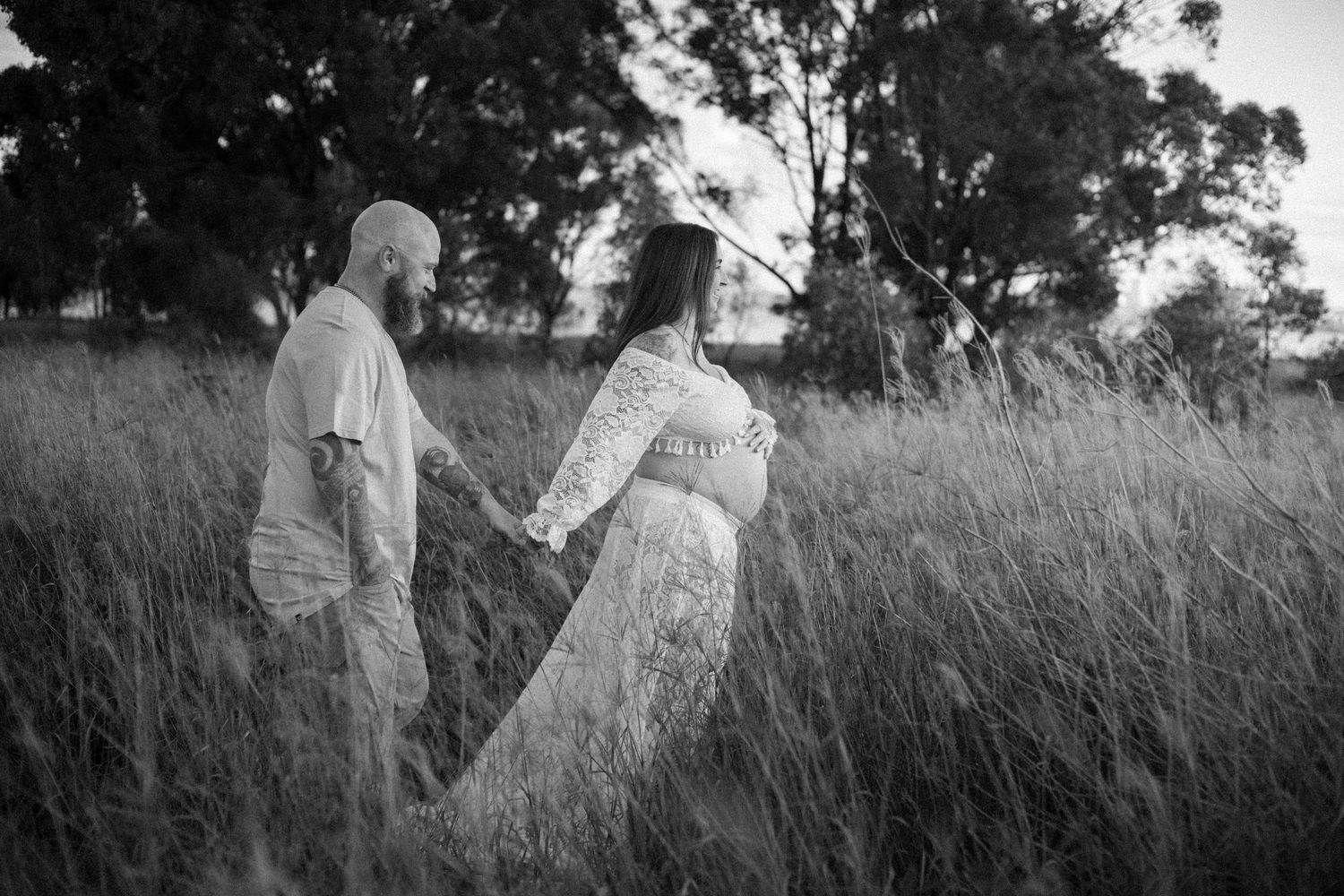 Black and white photo of a couple holding hands in tall grass field, woman in lace maternity gown, trees in background.