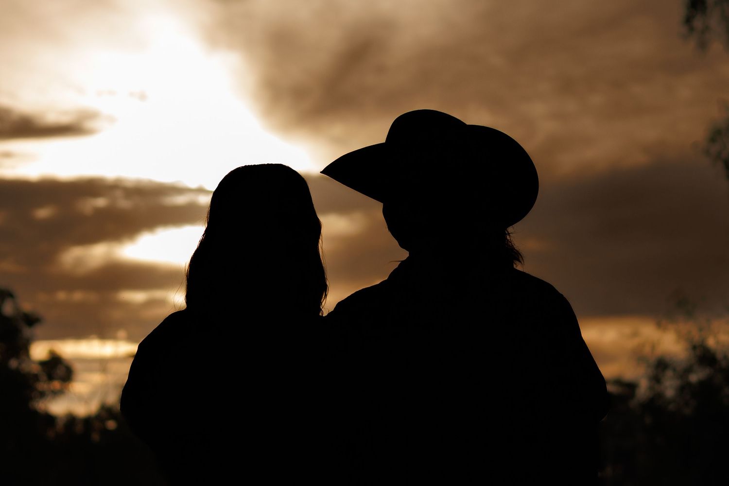 Silhouette of a couple at sunset, one wearing a cowboy hat, against a dramatic cloudy sky.