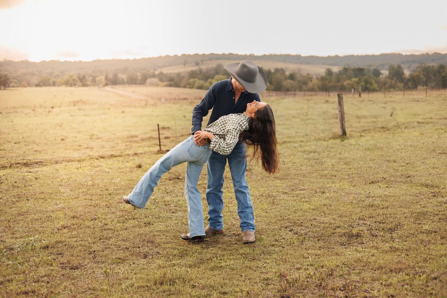 Couple dancing in a sunlit green field, man dipping woman in jeans and cowboy hat with rolling hills behind.