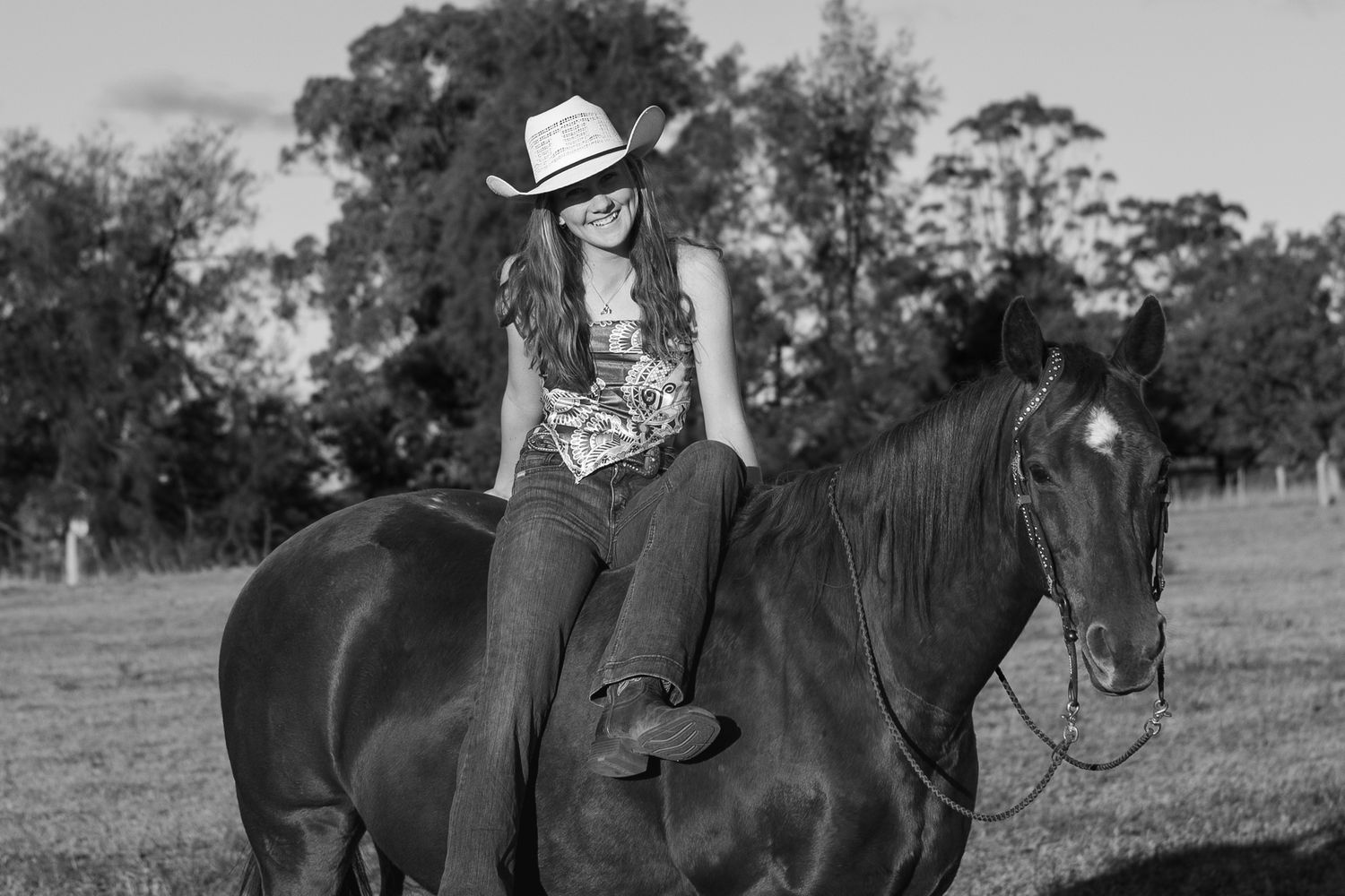 A young woman in a cowboy hat rides a dark horse in a grassy field with trees in the background.
