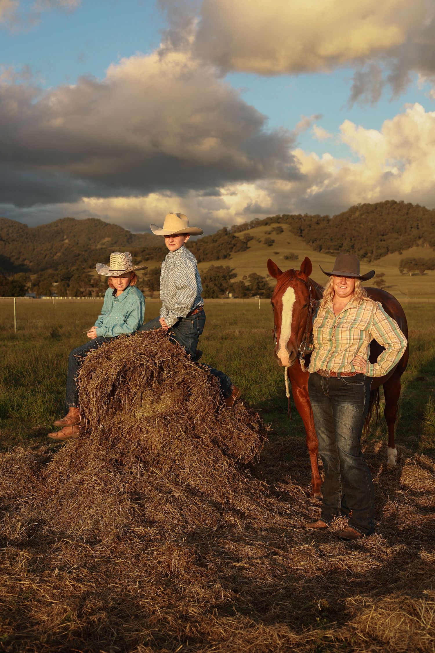 Three cowboys in hats pose with a chestnut horse by a hay bale in a green pasture at golden hour.