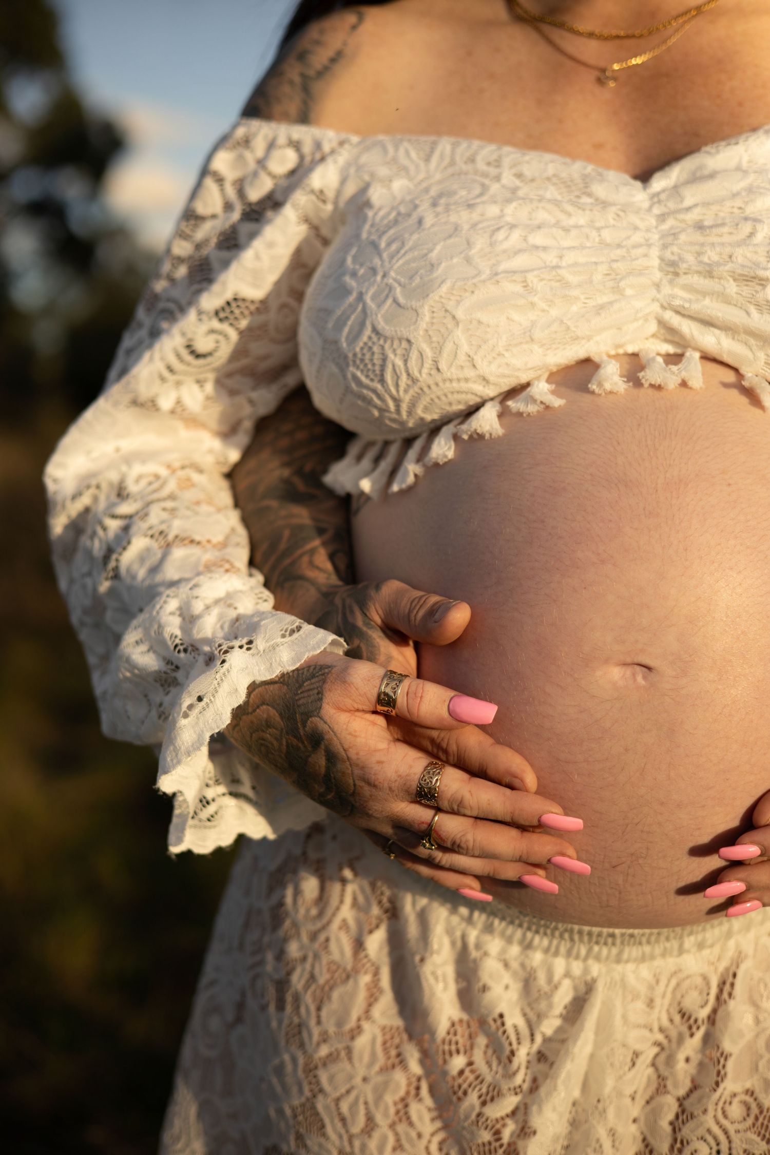 Pregnant woman in white lace off-shoulder dress cradling baby bump, wearing silver rings with pink manicure outdoors.