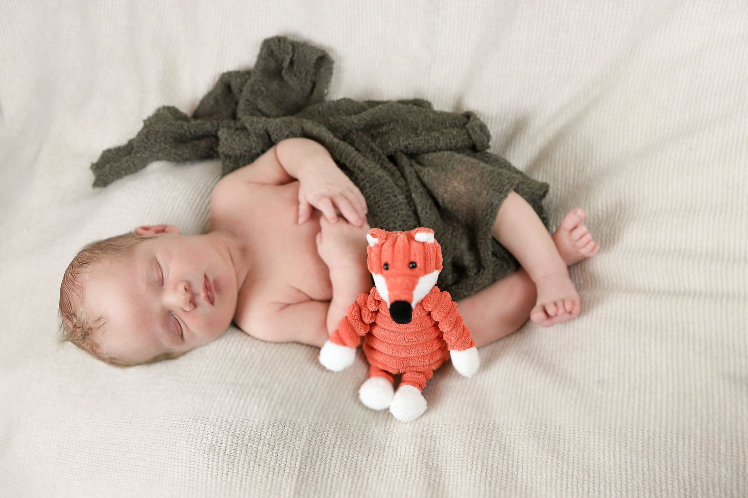 Sleeping baby with orange plush fox toy on white bedding.