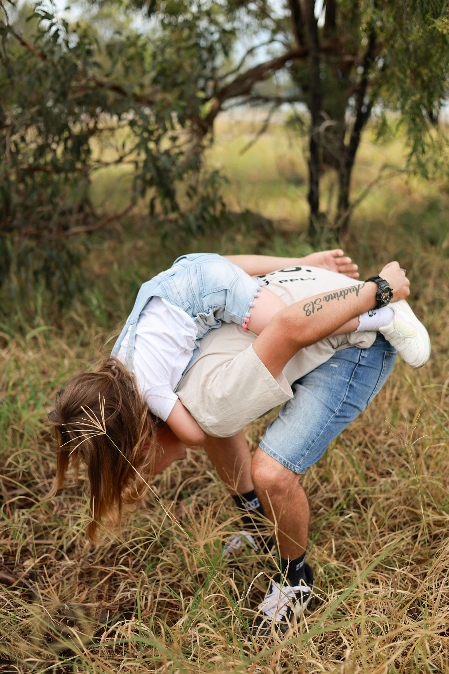 A flexible person bending forward touching the ground in a grassy outdoor field surrounded by trees.