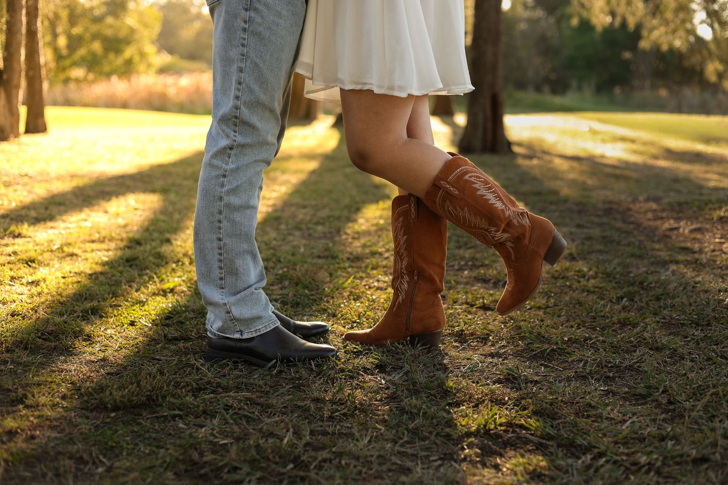 Couple standing close together outdoors, woman wearing brown cowboy boots and white dress, man in jeans and casual shoes.