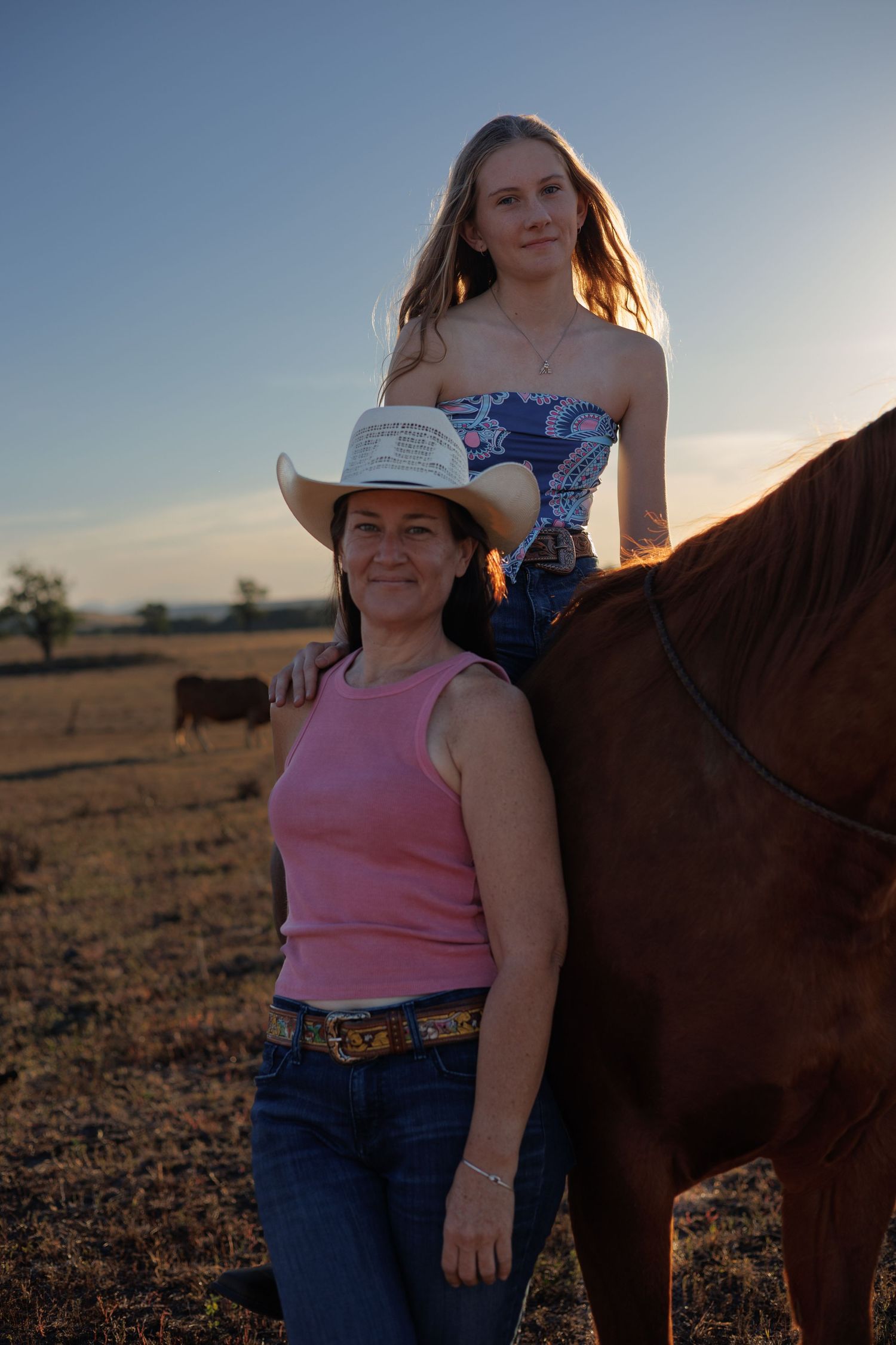 Two women pose with a brown horse at sunset in a rural field, one wearing a cowboy hat and pink tank top.