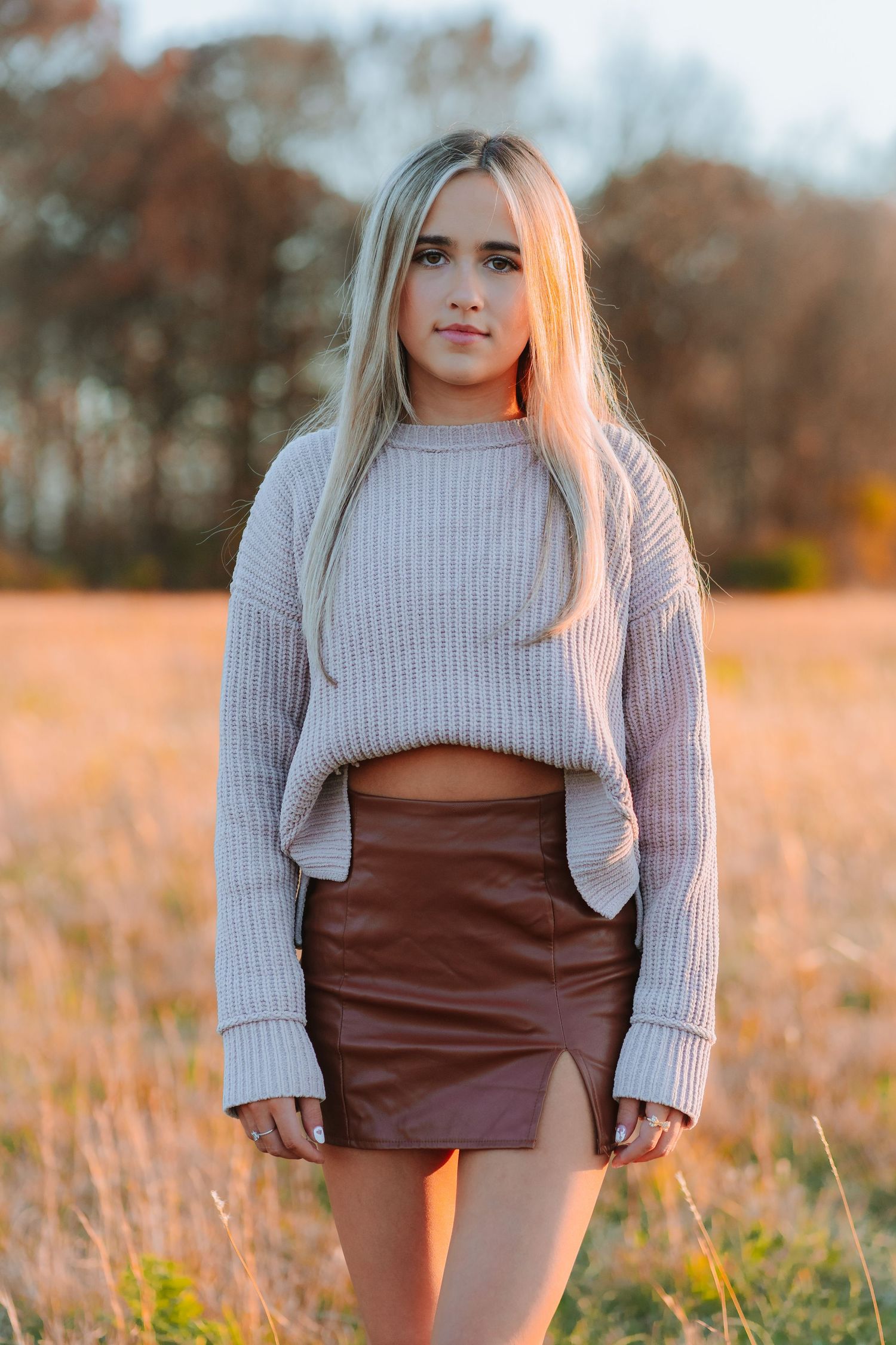 A fashionable fall outfit featuring a cropped grey sweater and brown leather mini skirt in a golden sunset field.
