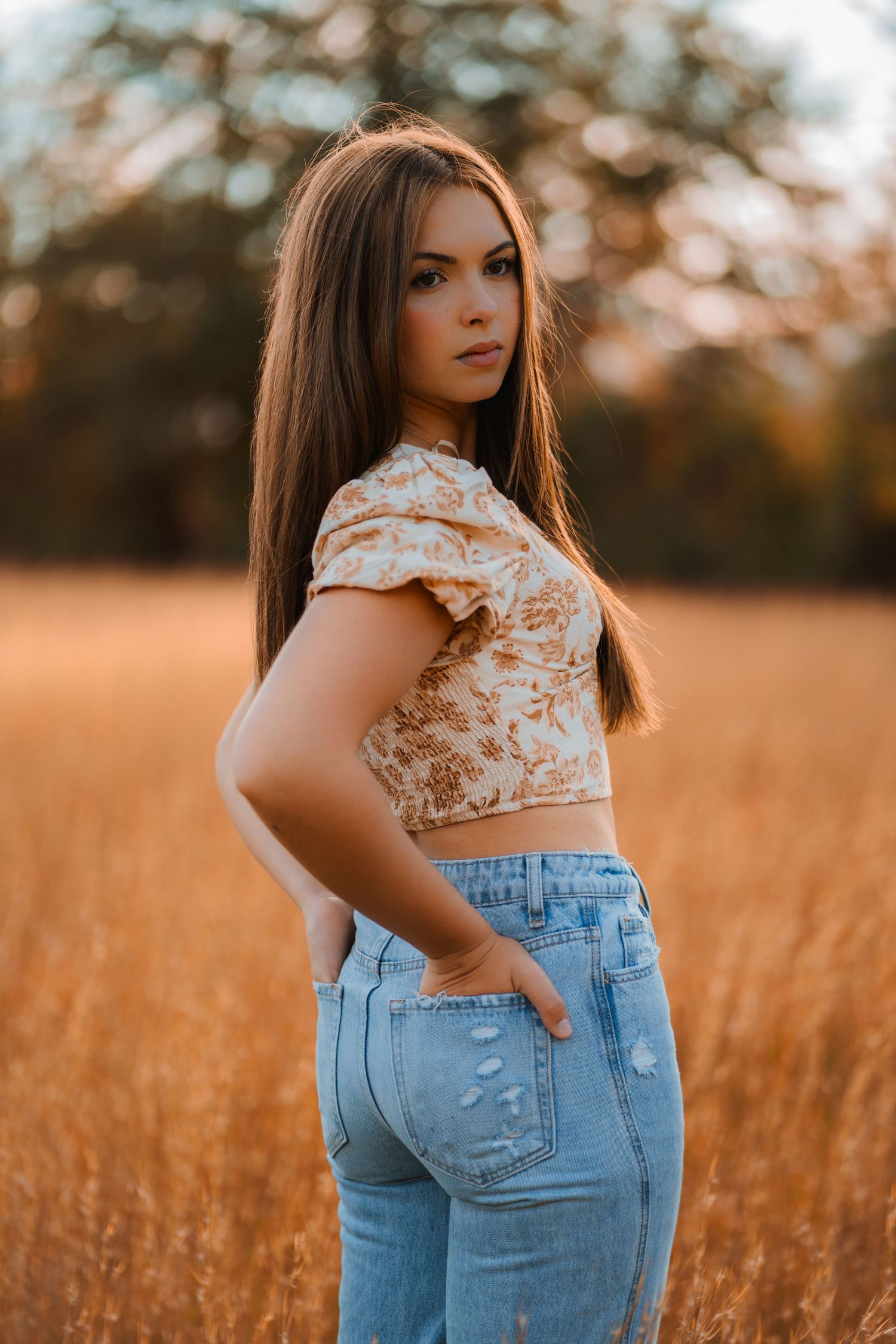 Portrait in a golden wheat field wearing light floral crop top and high-waisted jeans during sunset.