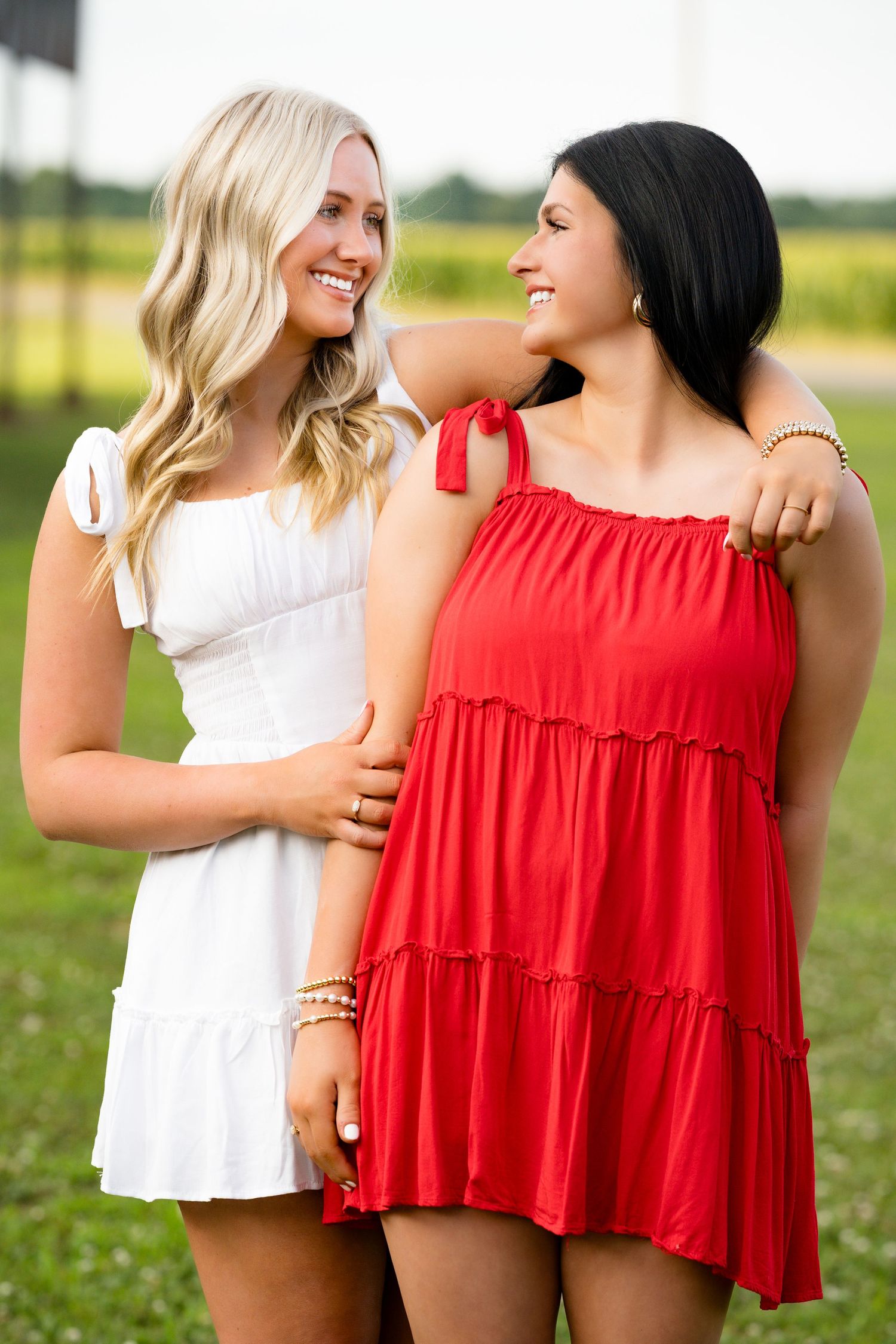 Two friends laugh together wearing contrasting white and red ruffled summer tops in a sunny outdoor field.