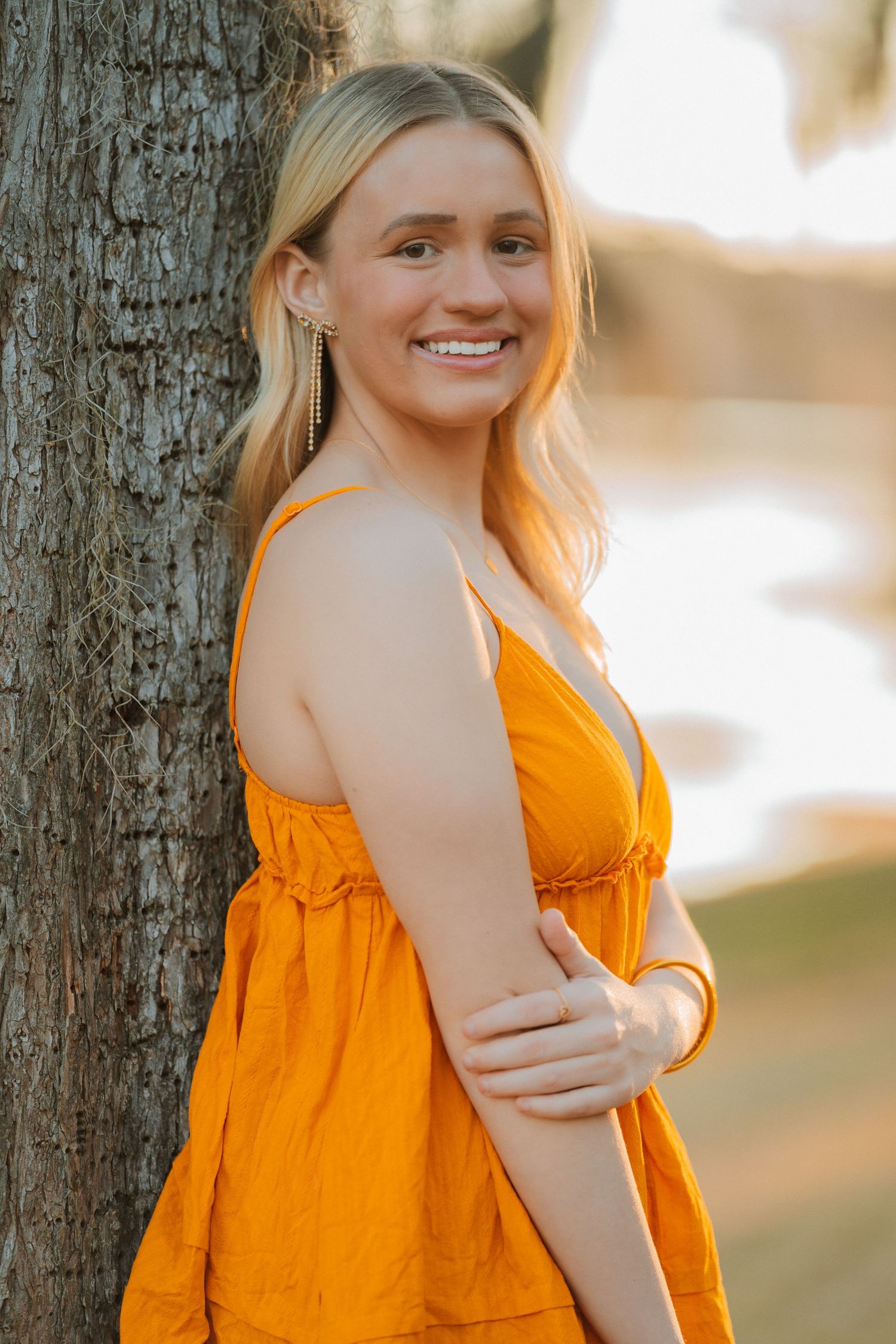 A person in a bright orange sundress leans against a tree trunk while smiling during golden hour lighting.