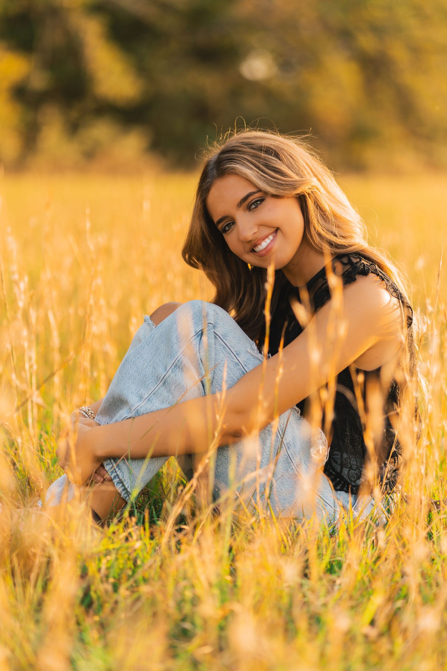 A smiling figure sits in a golden wheat field at sunset wearing jeans and a black top.