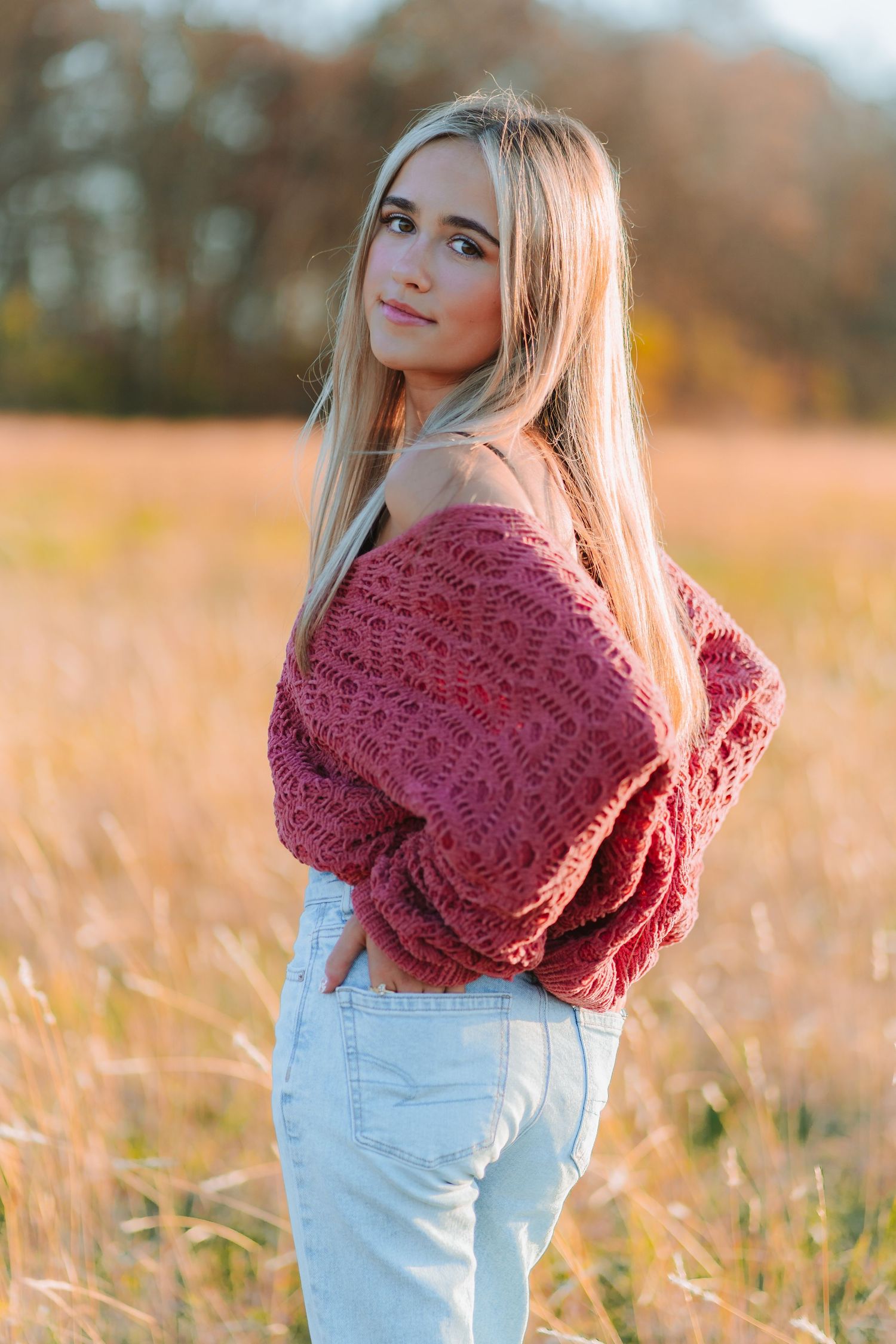 Long-haired woman in burgundy knit sweater and light jeans poses in golden autumn field at sunset.