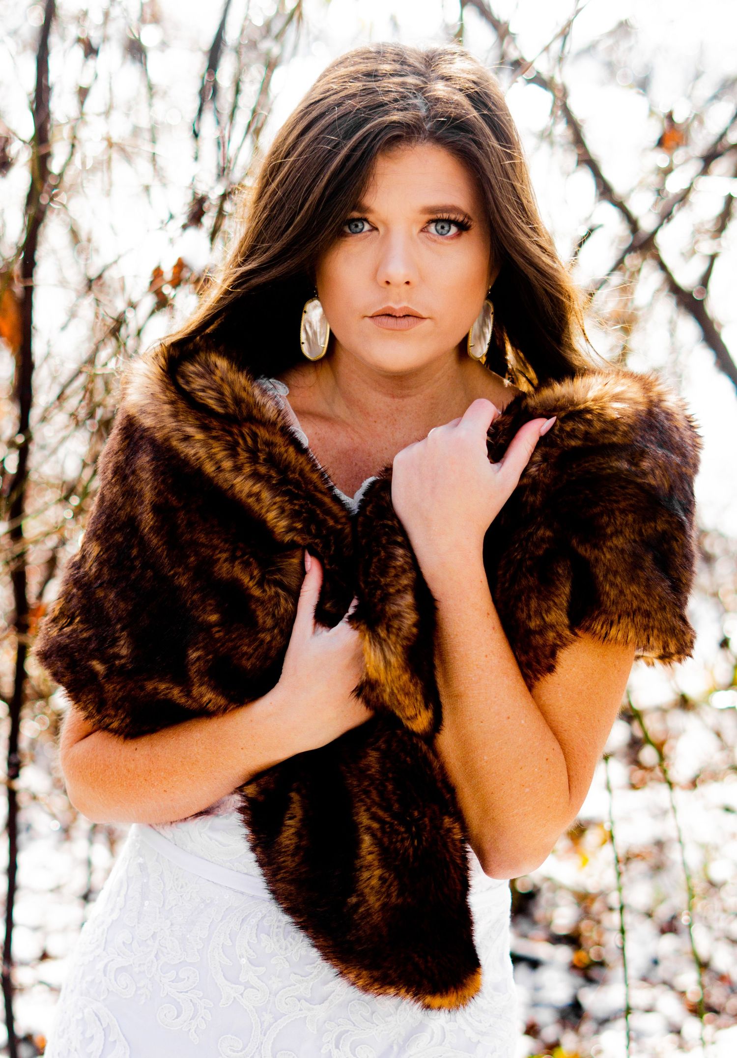 A woman in a white dress poses outdoors wearing a brown fur stole and silver earrings in an autumn setting.