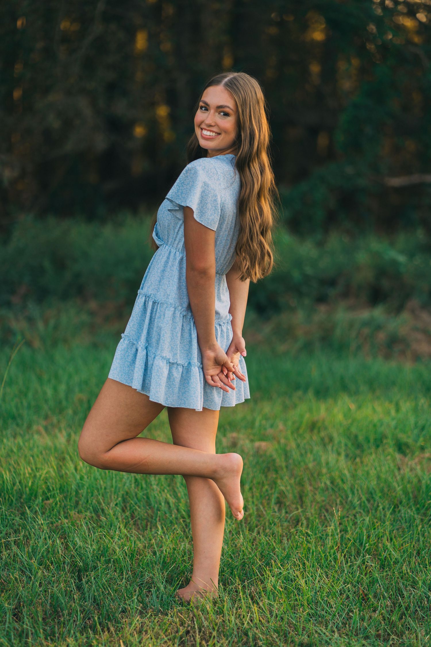 A person in a light blue tiered summer dress poses playfully while standing barefoot in a grassy field at sunset.