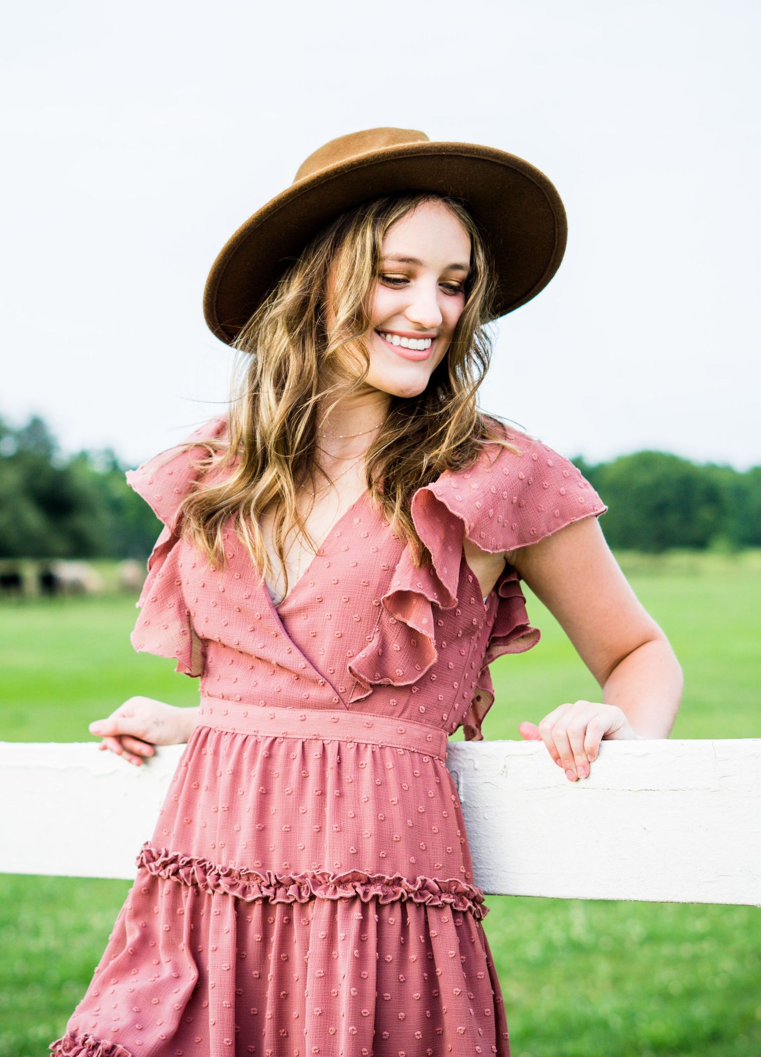 Woman in pink ruffled dress and brown hat leaning against white fence in sunny countryside field.