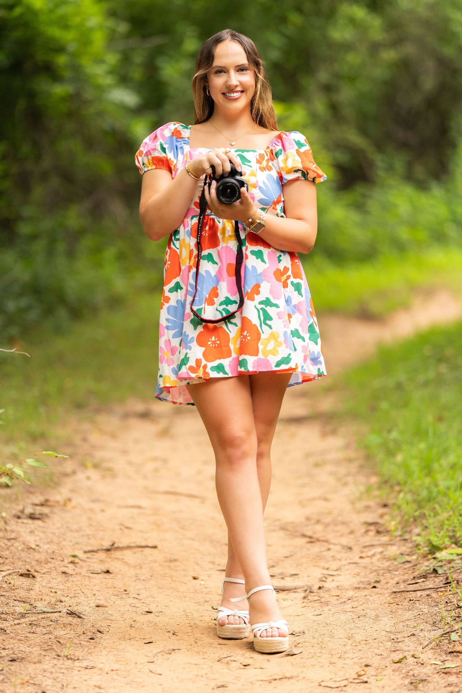 Someone wearing a colorful floral print off-shoulder dress poses outdoors on a dirt path surrounded by greenery.