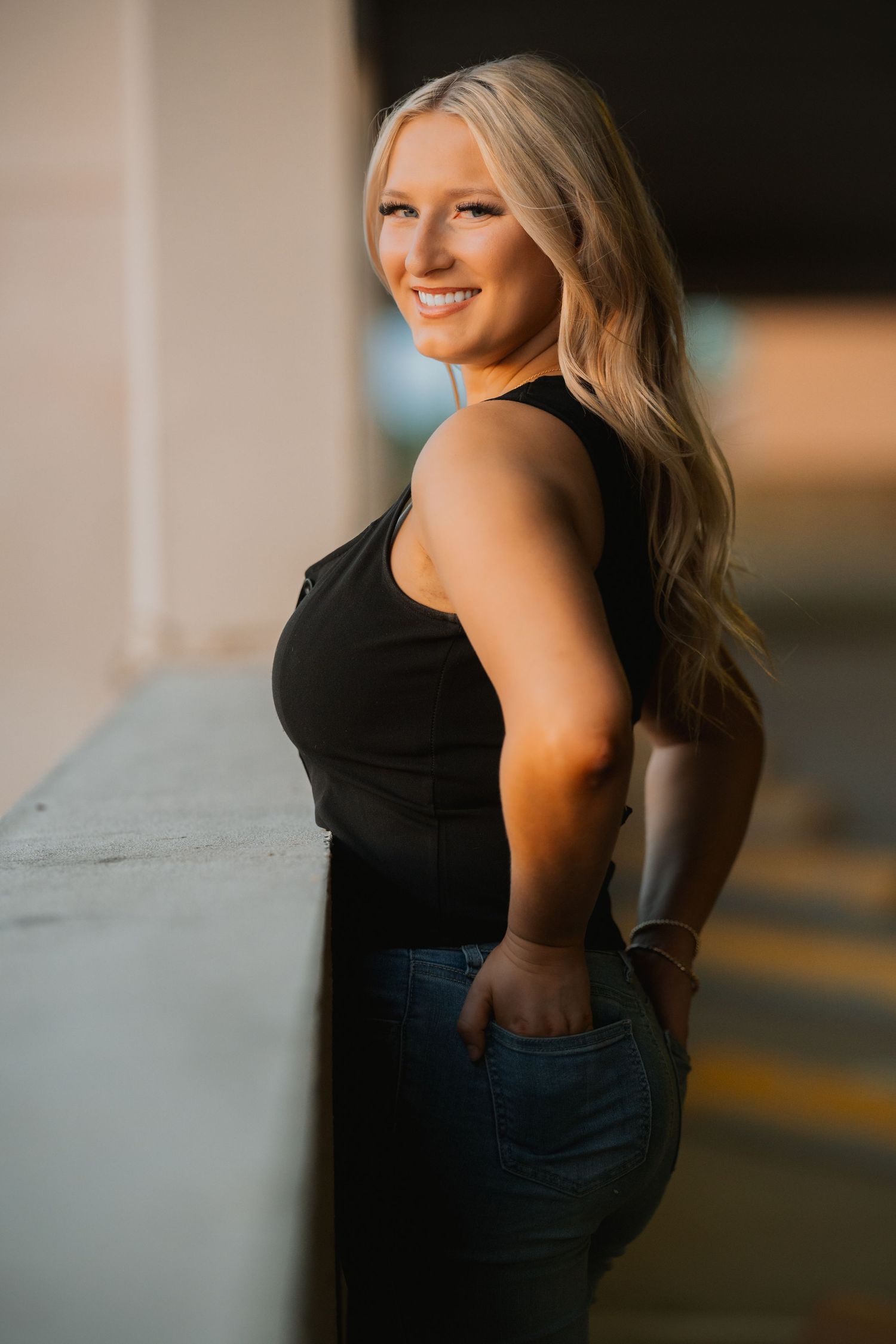 A portrait of someone in a black sleeveless top and jeans posing in profile against a shadowy background at sunset.