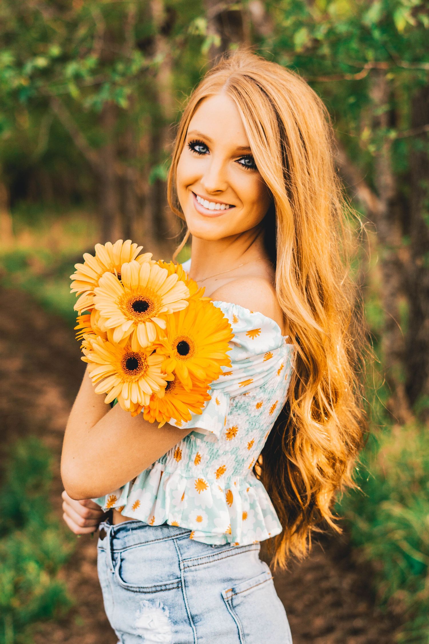 Young woman in floral top and denim holds vibrant yellow gerbera daisies while posing outdoors in natural setting.