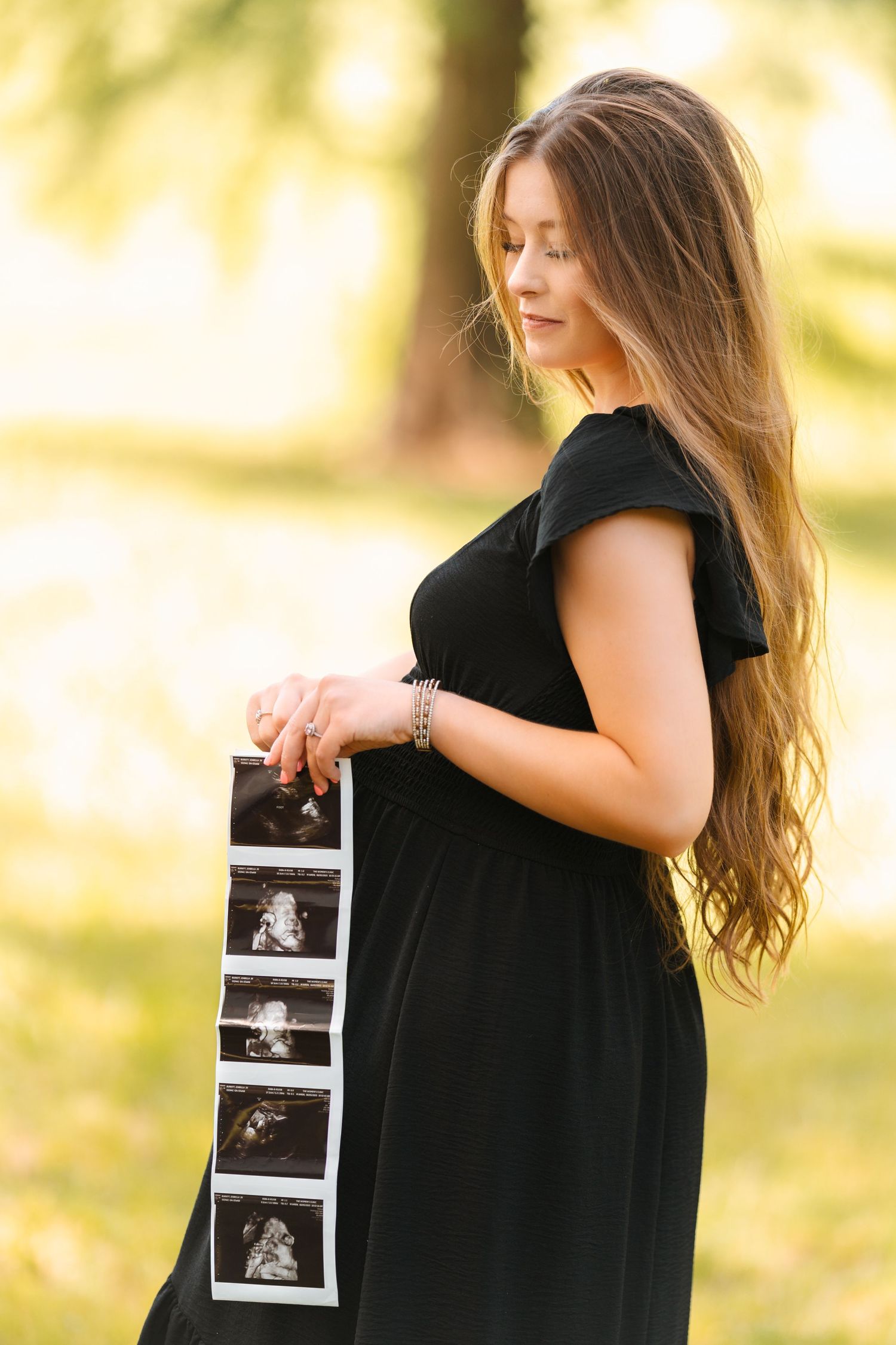 A pregnant woman in a black dress holds ultrasound photos in a sunlit outdoor setting with a soft bokeh background.
