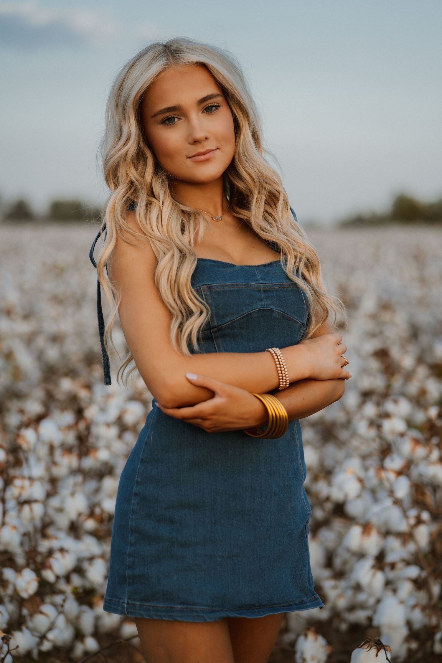 A portrait in a cotton field at sunset features someone in a denim dress with long blonde wavy hair.