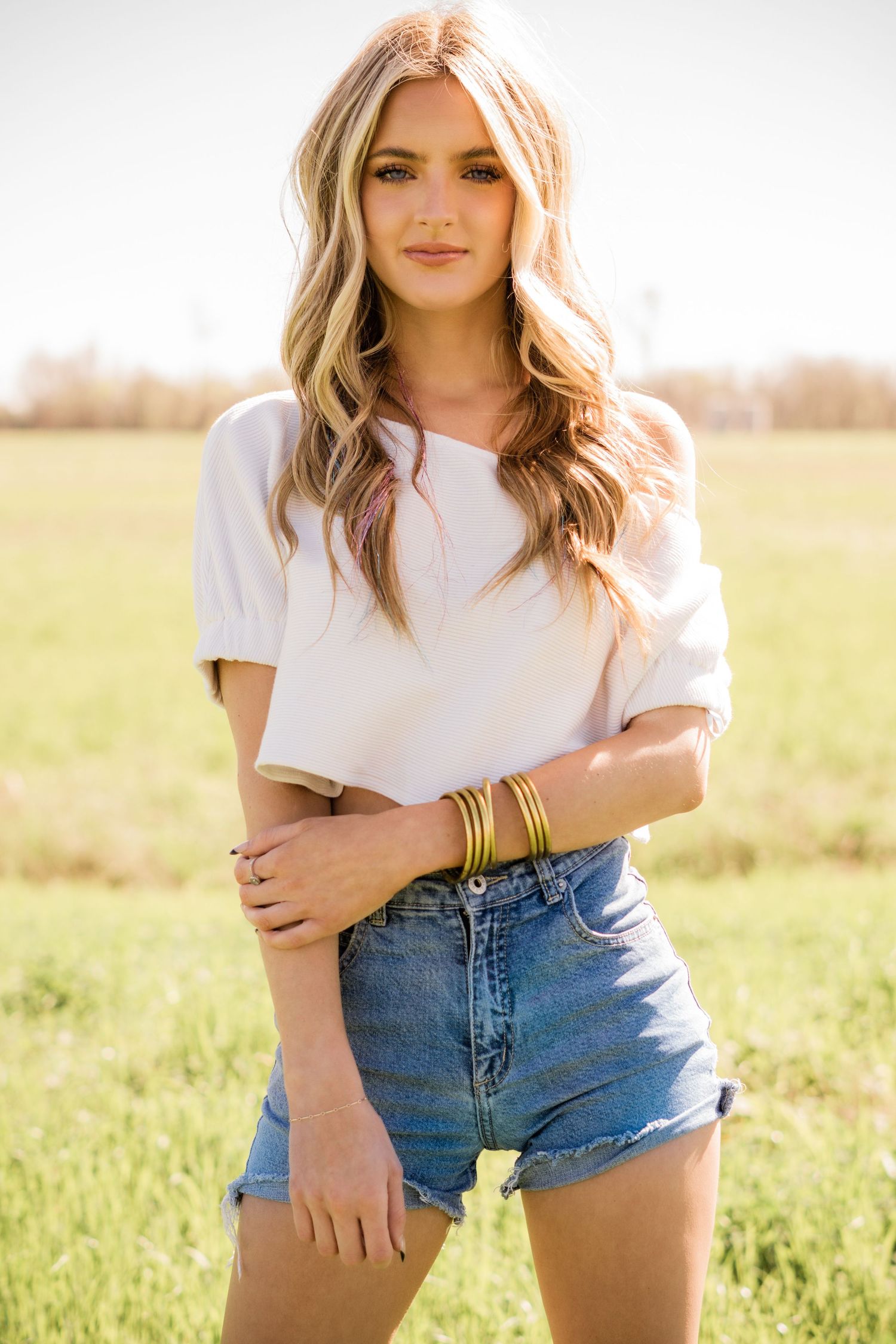 Young woman in white crop top and denim shorts poses in sunny field during golden hour.