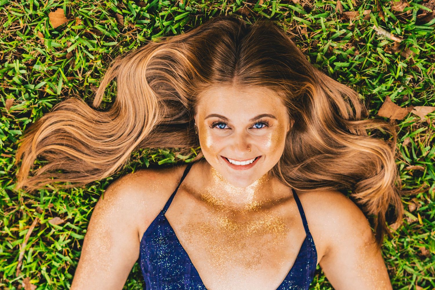 A person with long wavy blonde hair lies on green grass wearing a navy blue top while smiling.
