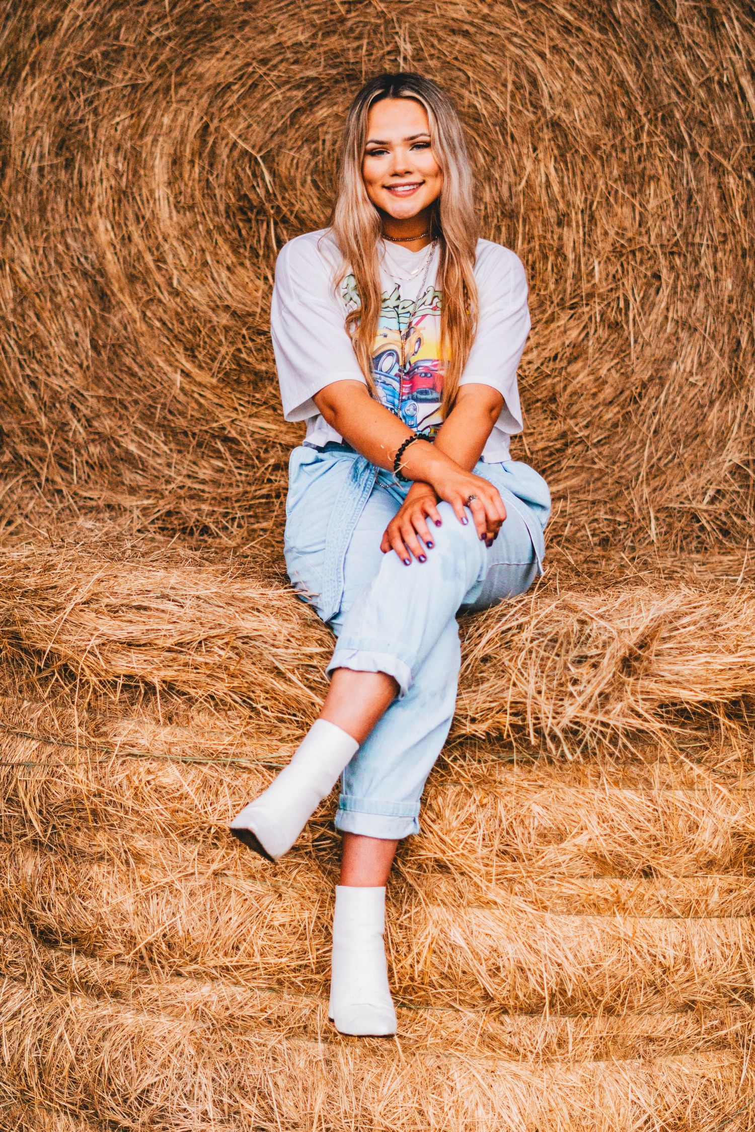 A person in casual wear sits on a hay bale, wearing a graphic t-shirt and light denim jeans with white socks.