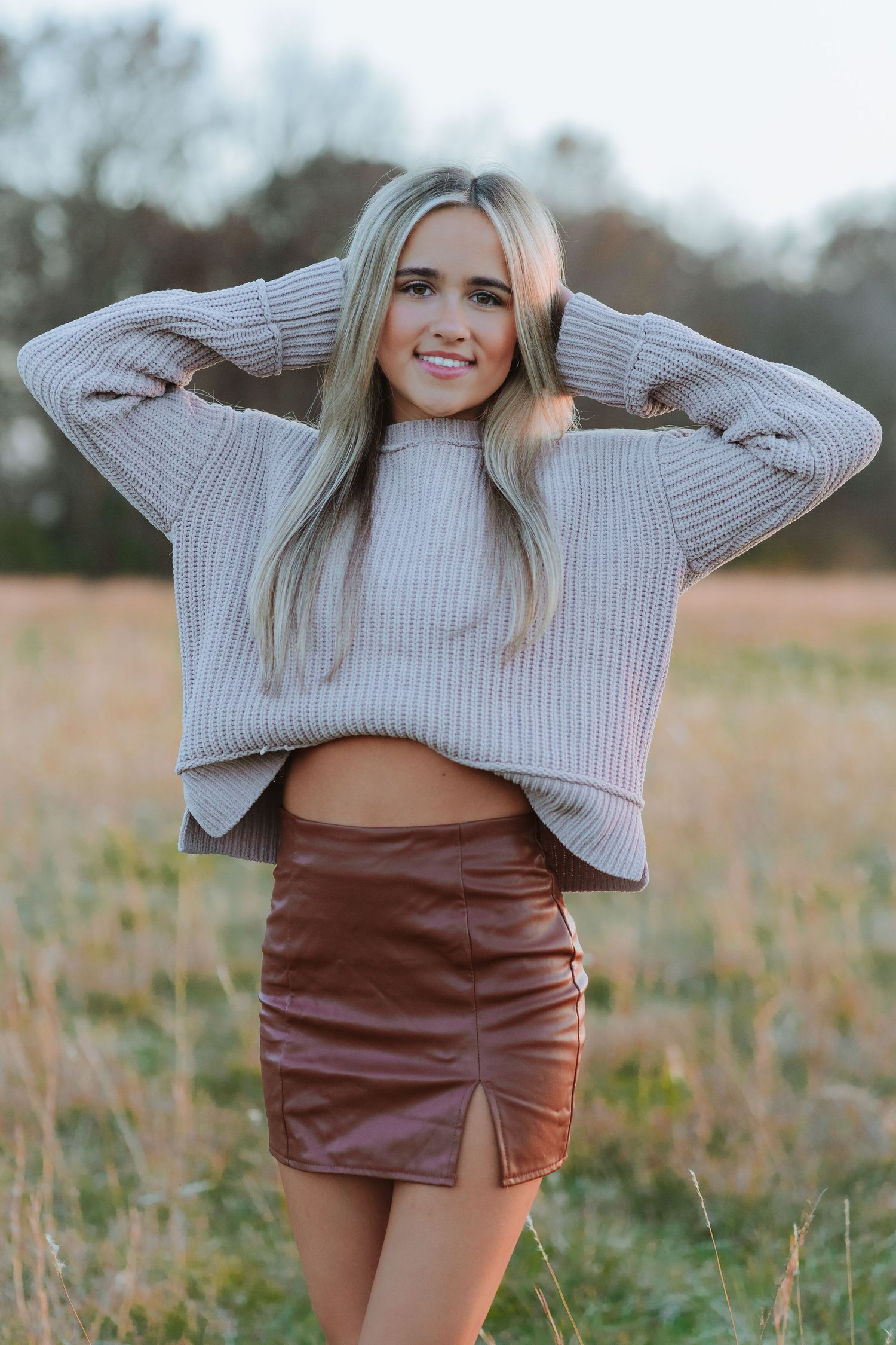 A cozy beige knit sweater paired with a brown leather mini skirt in an autumn field at sunset.