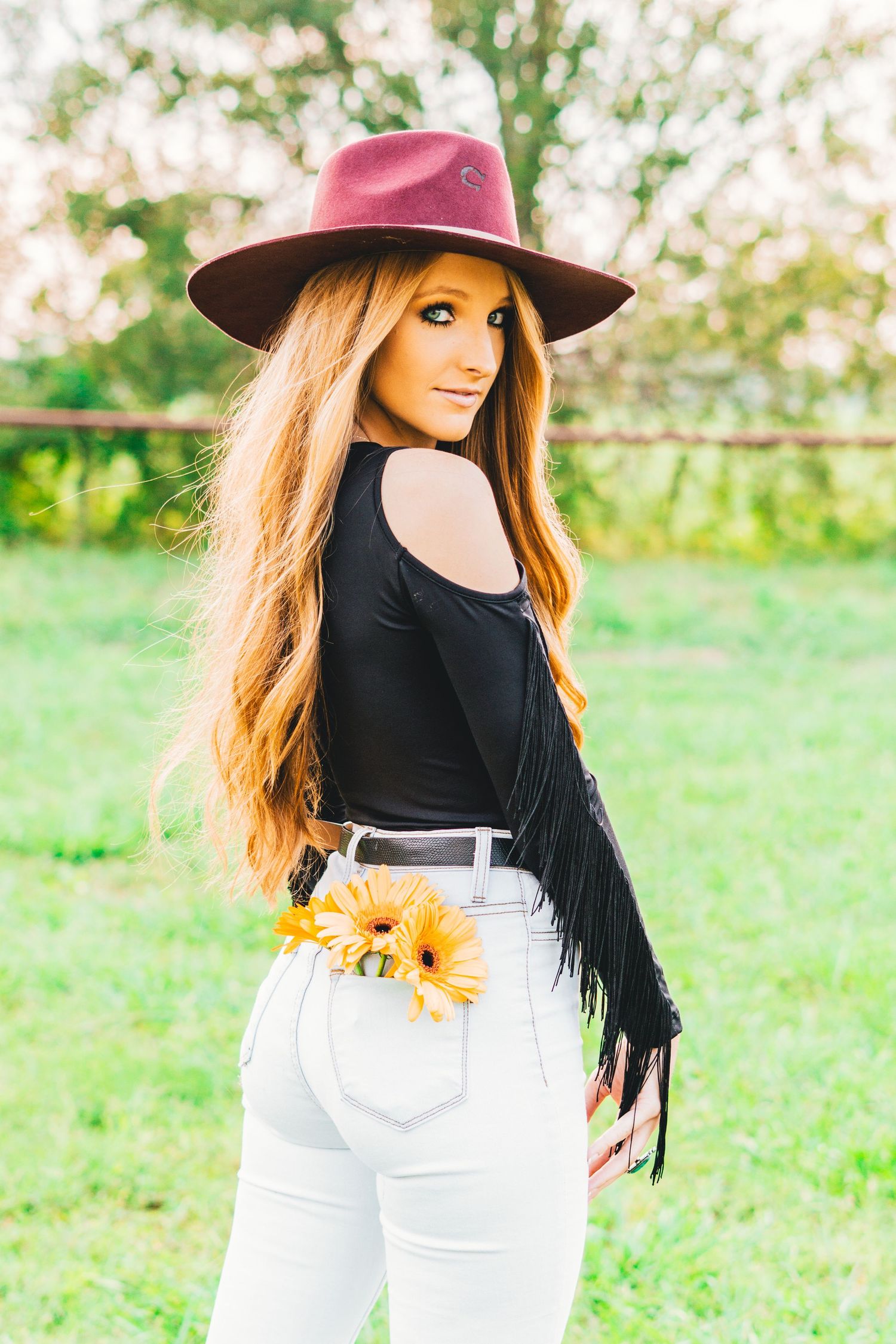 Stylish western outfit with black fringe top, white jeans, burgundy hat and sunflowers in back pocket in outdoor setting.