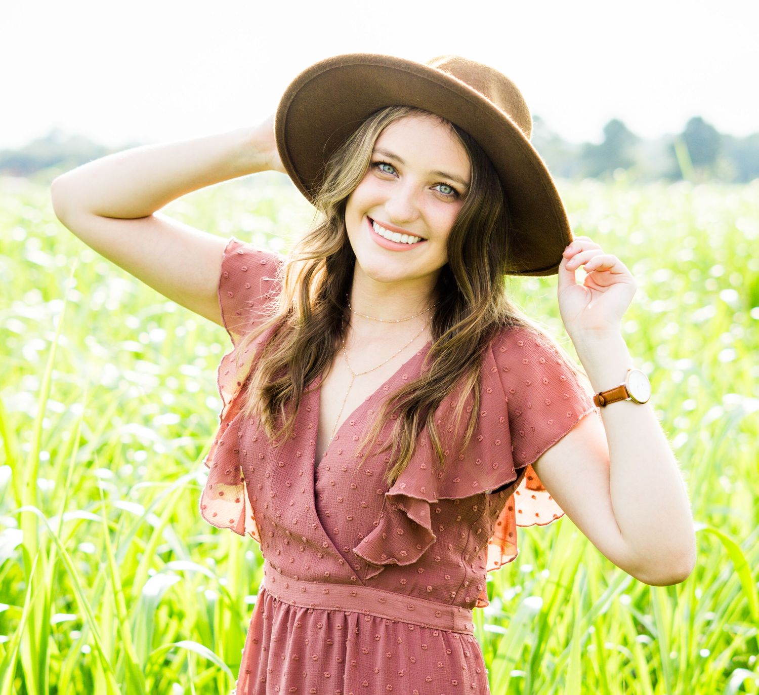 A person in a wide brimmed hat and dusty rose dress poses cheerfully in a sunny wildflower meadow.