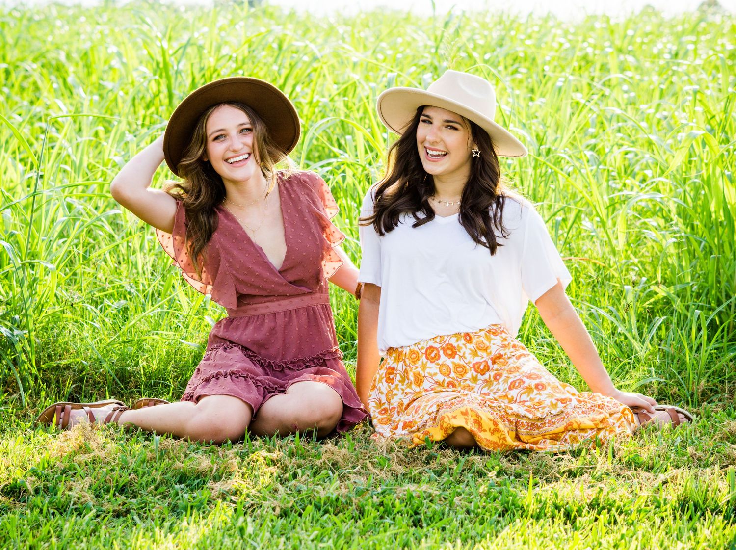 Two women in straw hats enjoying a sunny day outdoors while sitting on grass in colorful summer dresses.