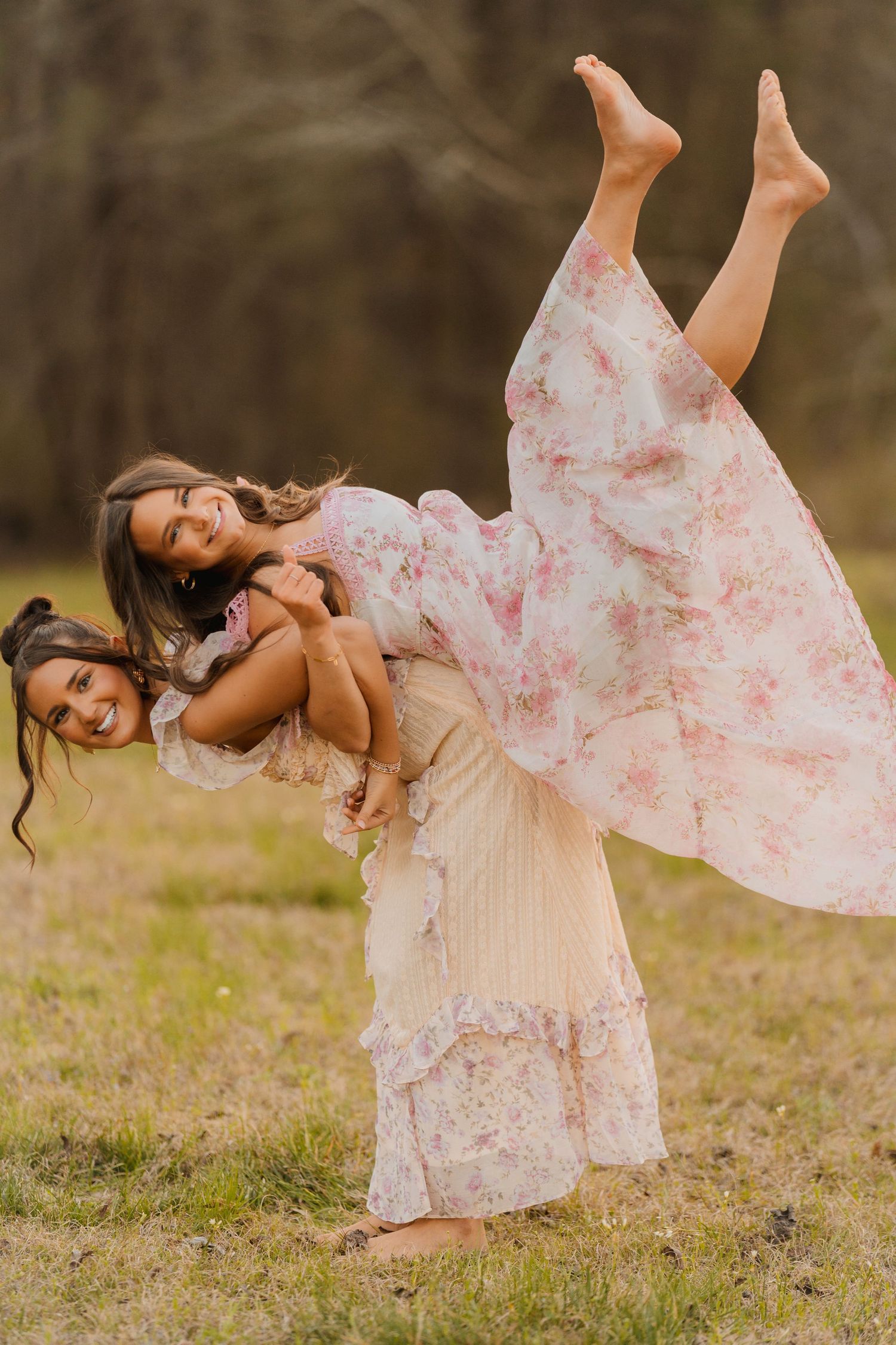 Two children in flowing floral dresses play together outdoors with one lifted in a playful pose against a blurred natural backdrop.