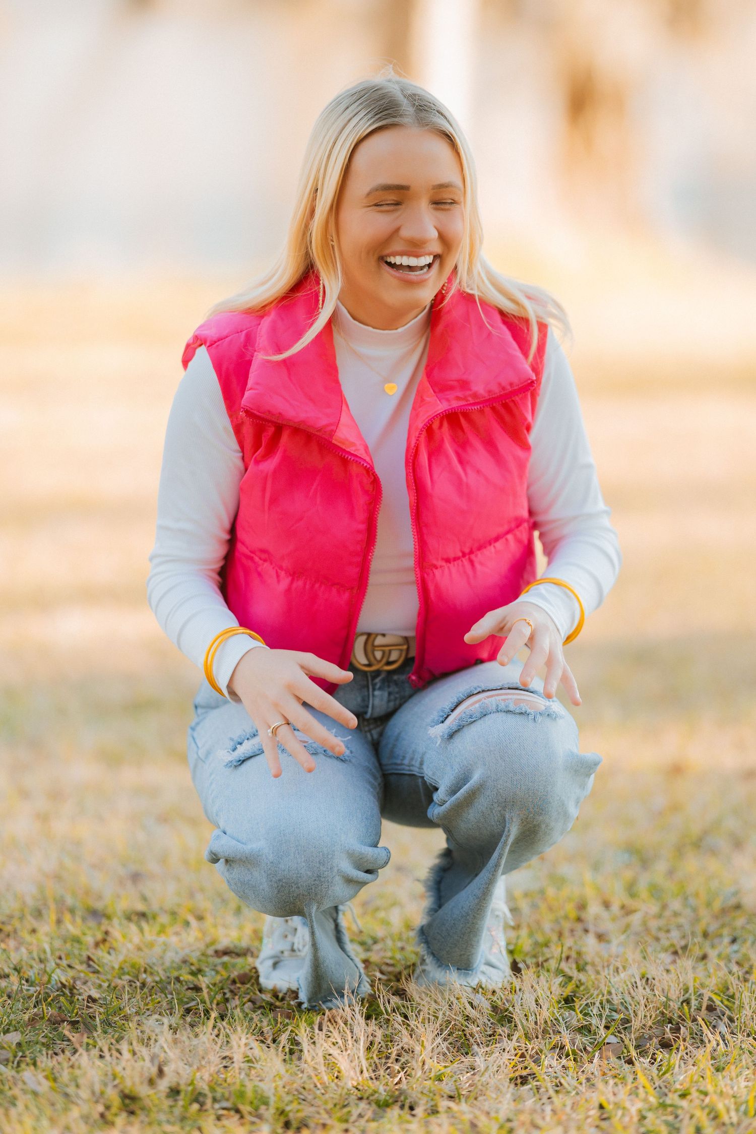 Casual outdoor portrait of someone crouching and smiling while wearing a bright pink puffer vest and light jeans.