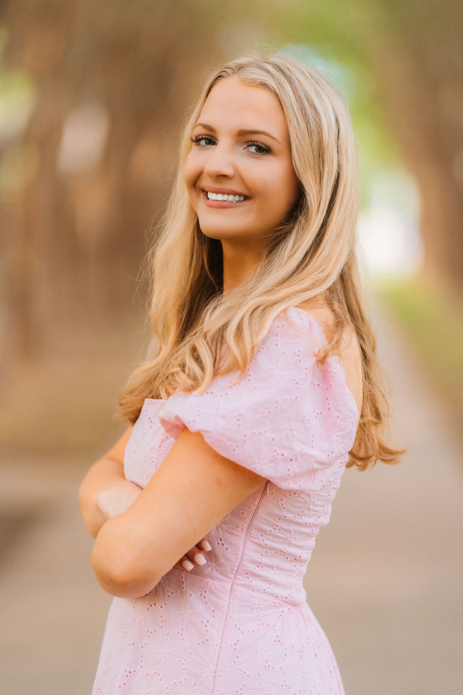 A portrait of someone wearing a pink lace dress with long blonde hair flowing down and posing with a warm smile outdoors.