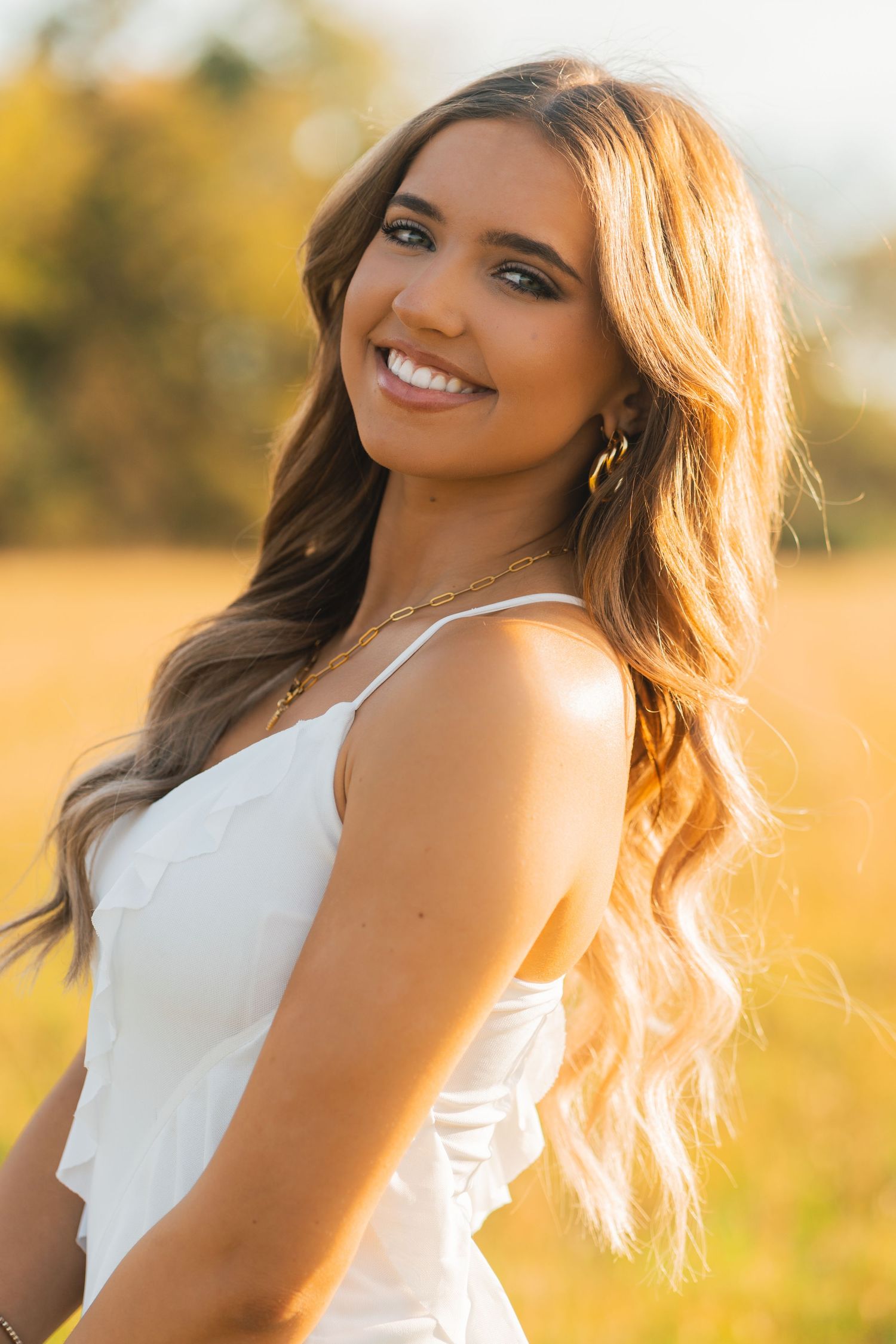 Portrait of someone in a white top smiling at sunset in a field with golden sunlight illuminating their wavy hair.