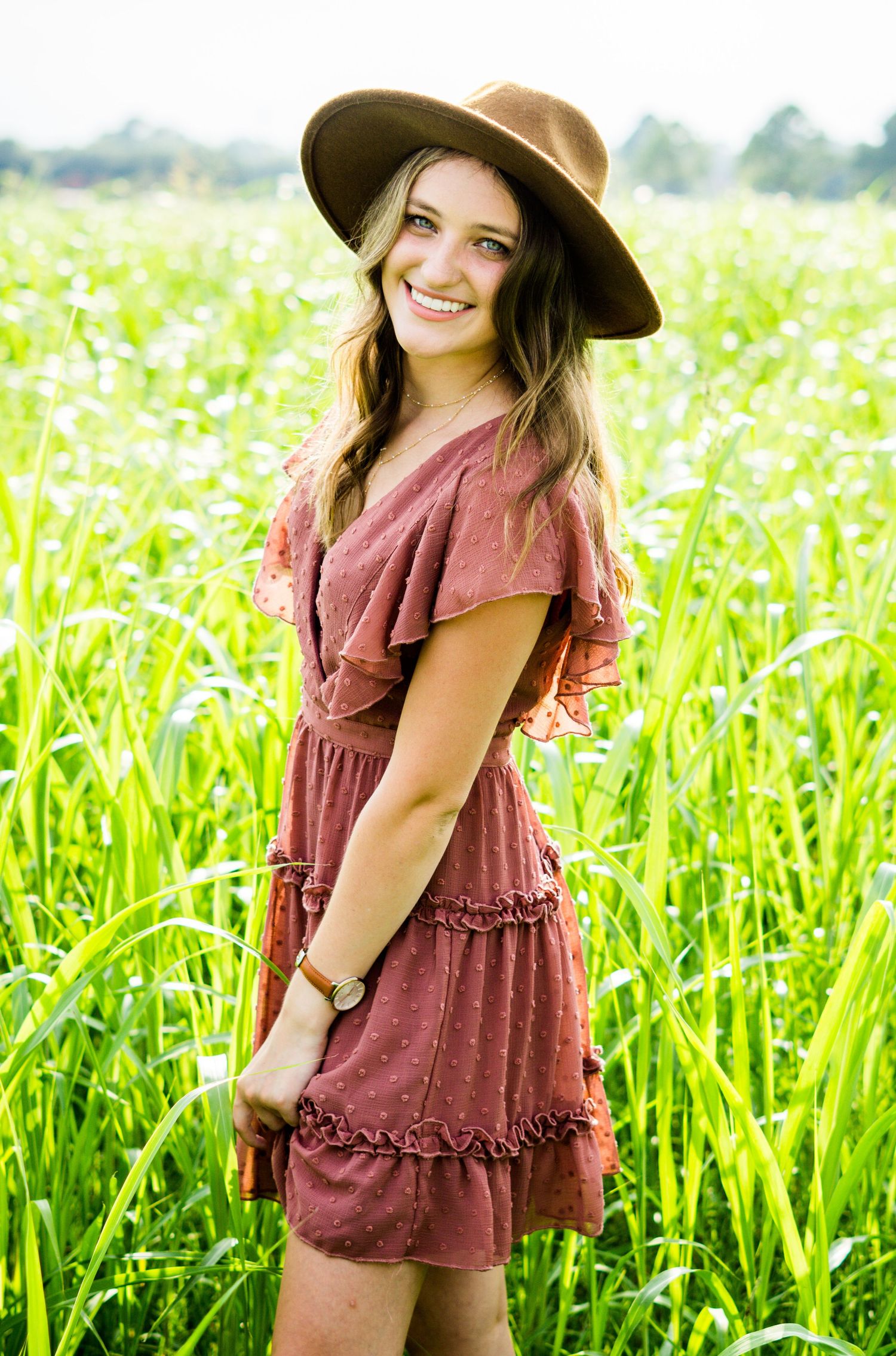 A person in a burgundy ruffle dress and wide-brimmed hat poses in a sunny field of tall green grass and wildflowers.