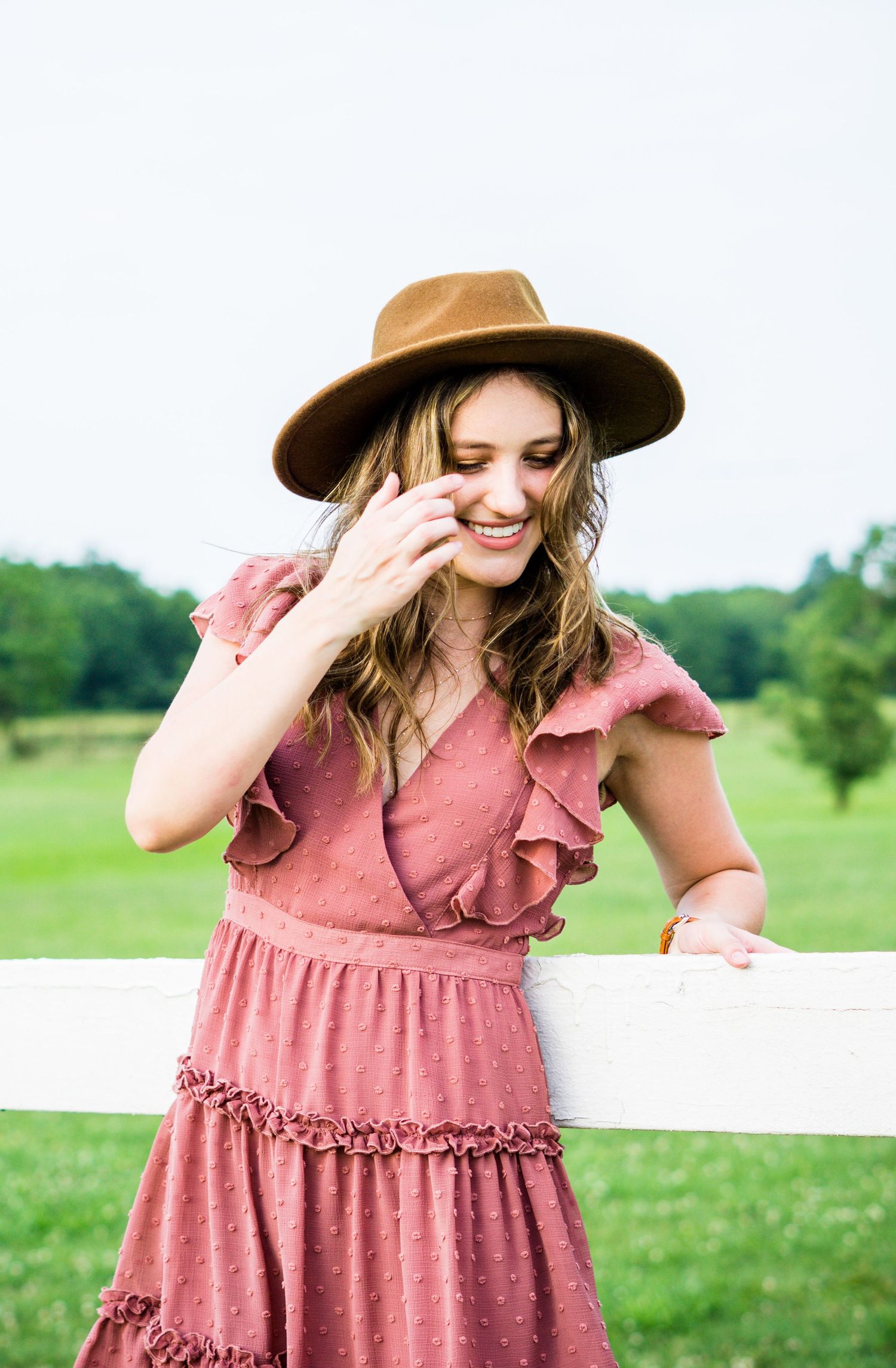 A person in a pink ruffled dress and brown hat leans against a white fence while laughing in a sunny field.