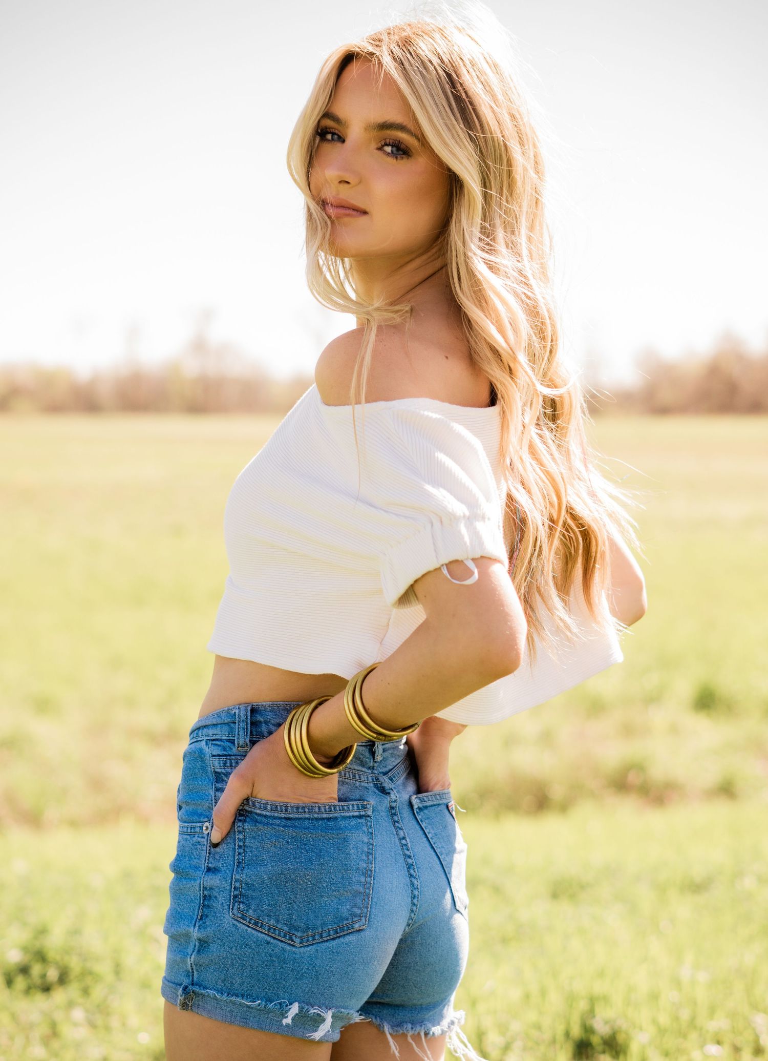 Person in white off-shoulder crop top and denim shorts poses in sunny field with golden light.