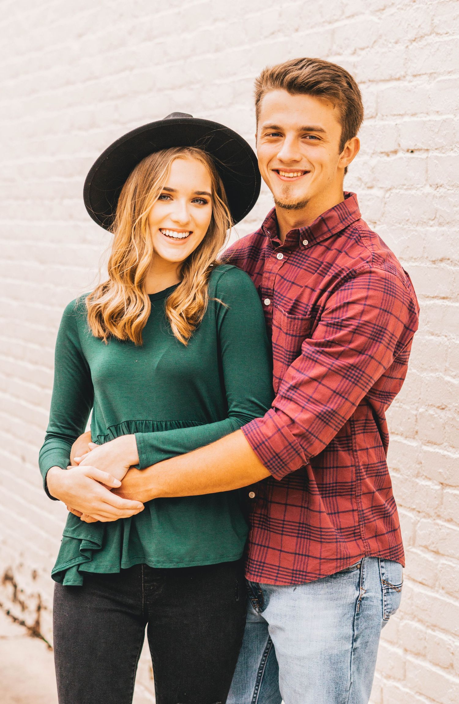 A couple poses together against a white brick wall, wearing a green sweater and plaid shirt.