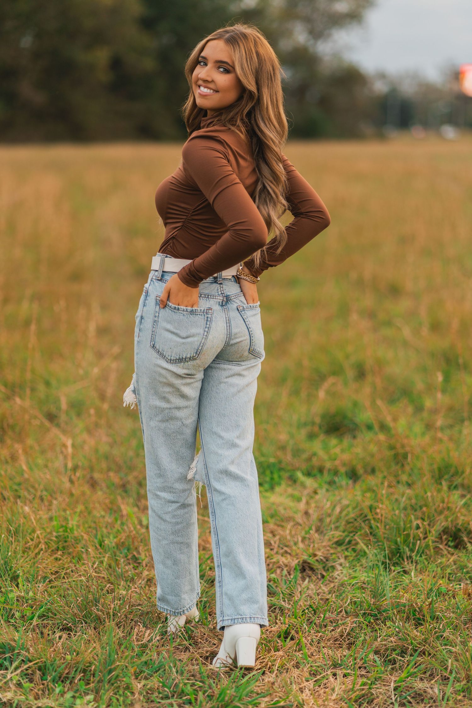 A person in a brown turtleneck and light wash mom jeans poses in a golden autumn field at sunset.
