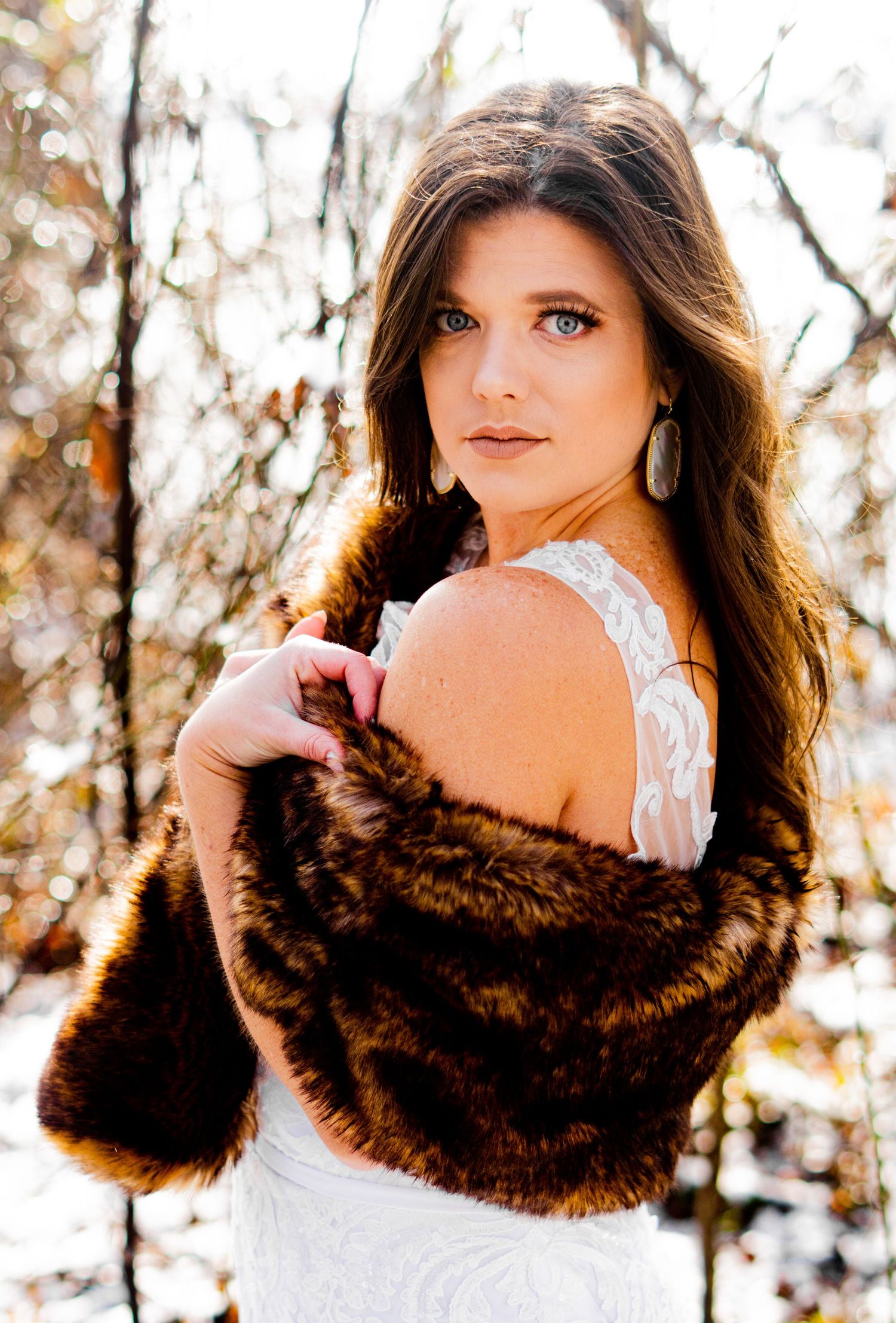 Woman in white dress and brown fur wrap poses in autumn woodland setting with warm sun glow through trees.
