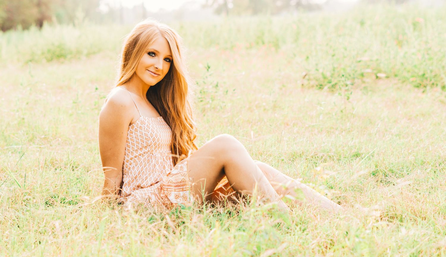 A person wearing a light pink dress sits casually in a sun-lit meadow with tall grass and soft lighting.