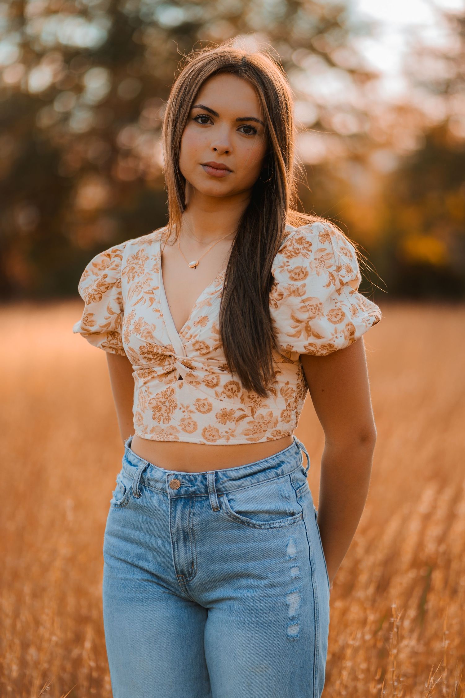 A person wearing a white floral crop top and high-waisted jeans poses in a golden field at sunset.