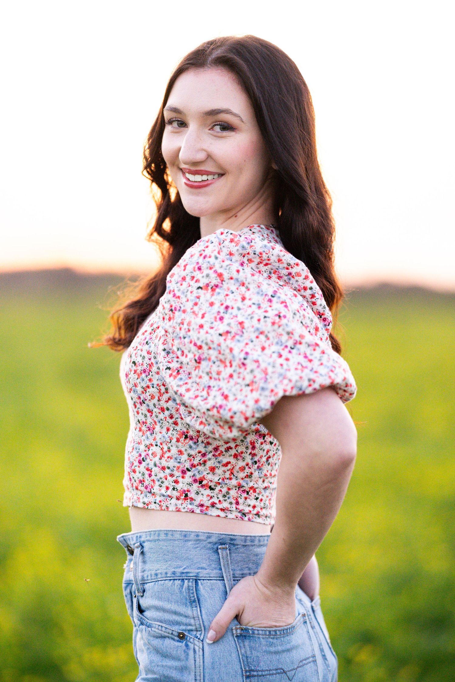 A person in a floral crop top and jeans poses in a field of yellow flowers against a sunset.