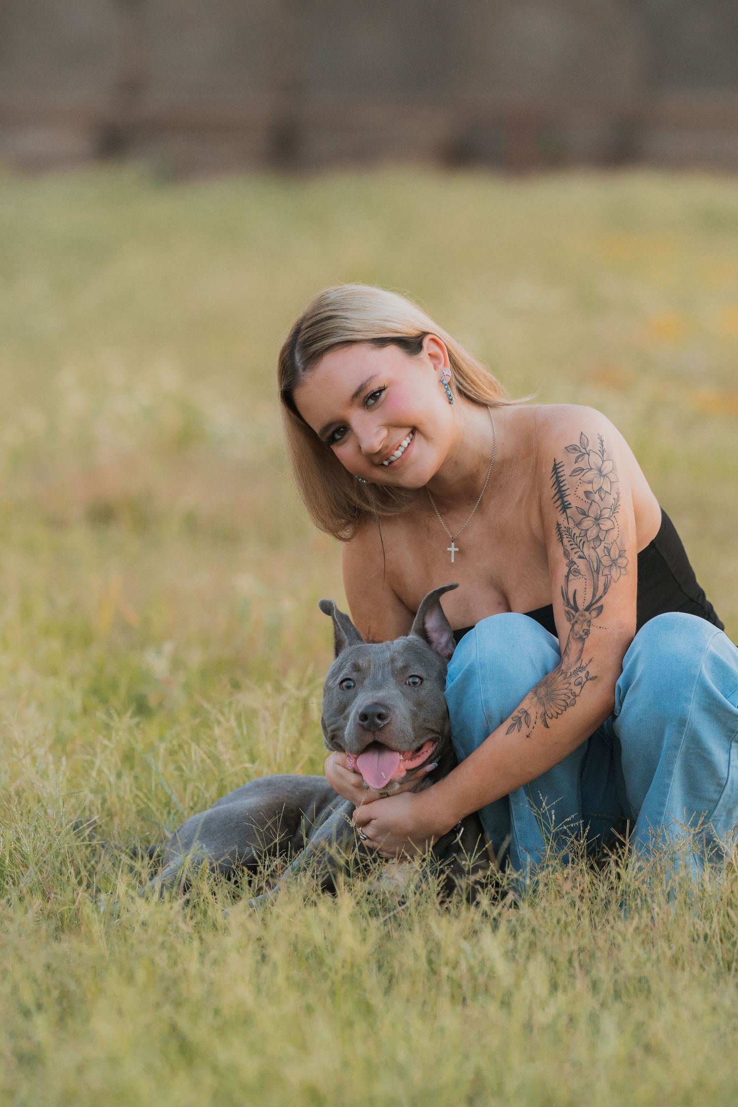 A person in blue jeans lies in green grass with a gray pitbull dog during an outdoor photoshoot.