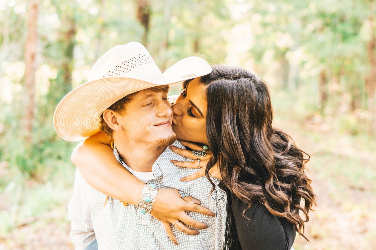 A couple shares a tender moment outdoors, with one wearing a cowboy hat as they embrace in soft natural lighting.
