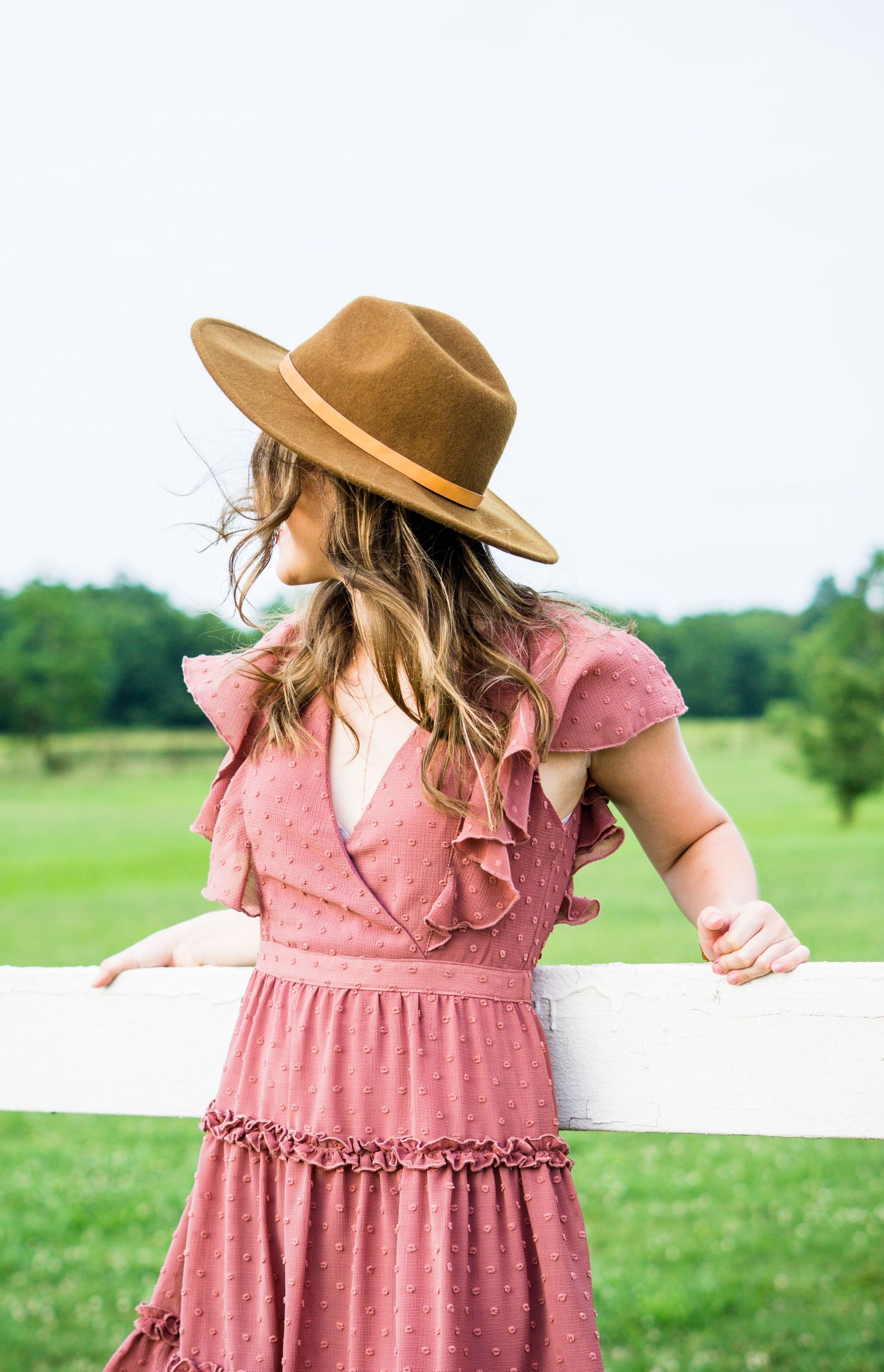 Woman in pink ruffled dress and brown fedora hat leans on white fence in countryside setting.